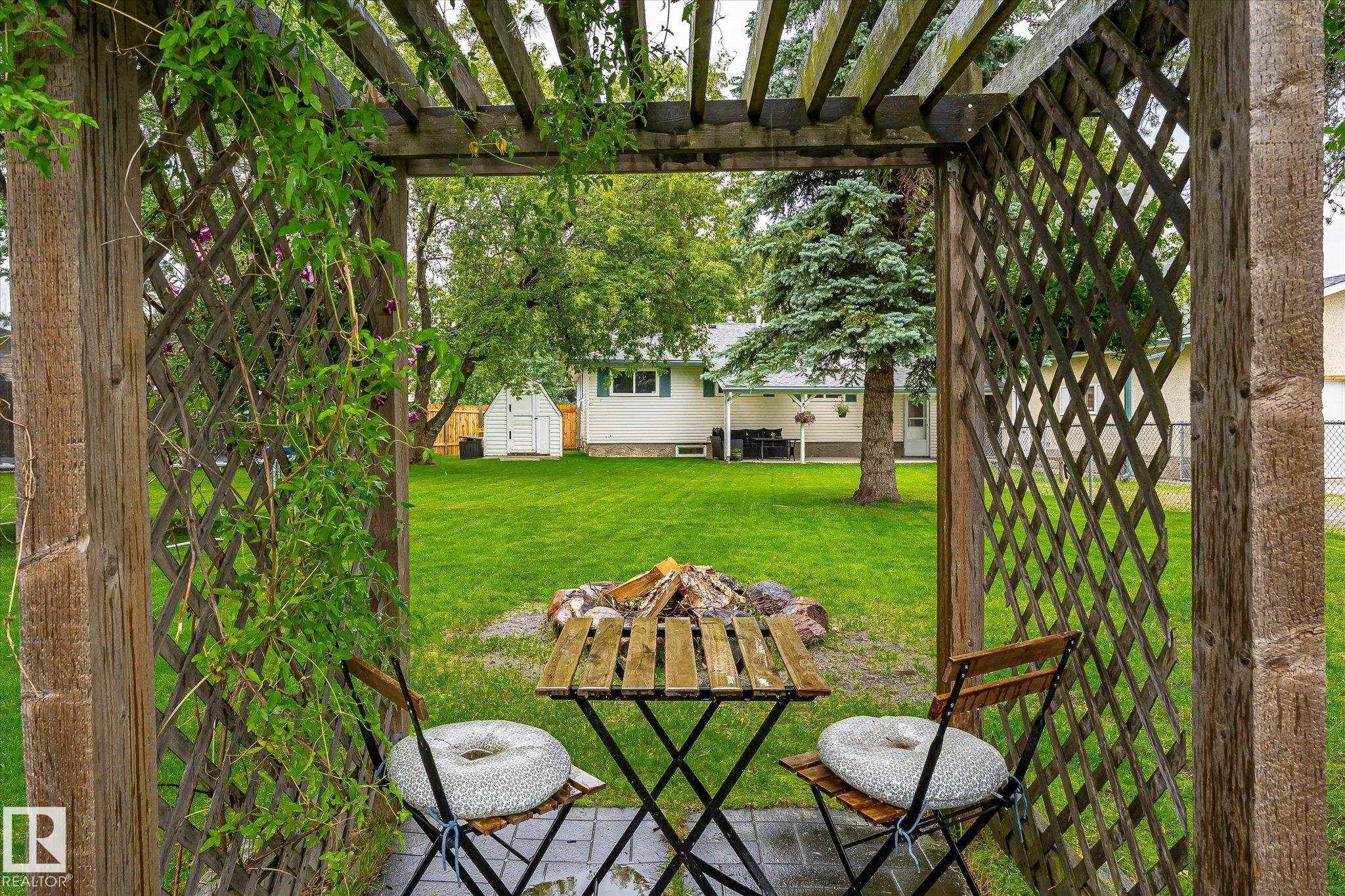 Wooden pergola with climbing greenery, paver patio, wood-finish table and chairs, and a fire pit area - 4921 47 Avenue, Legal, AB - Outdoor