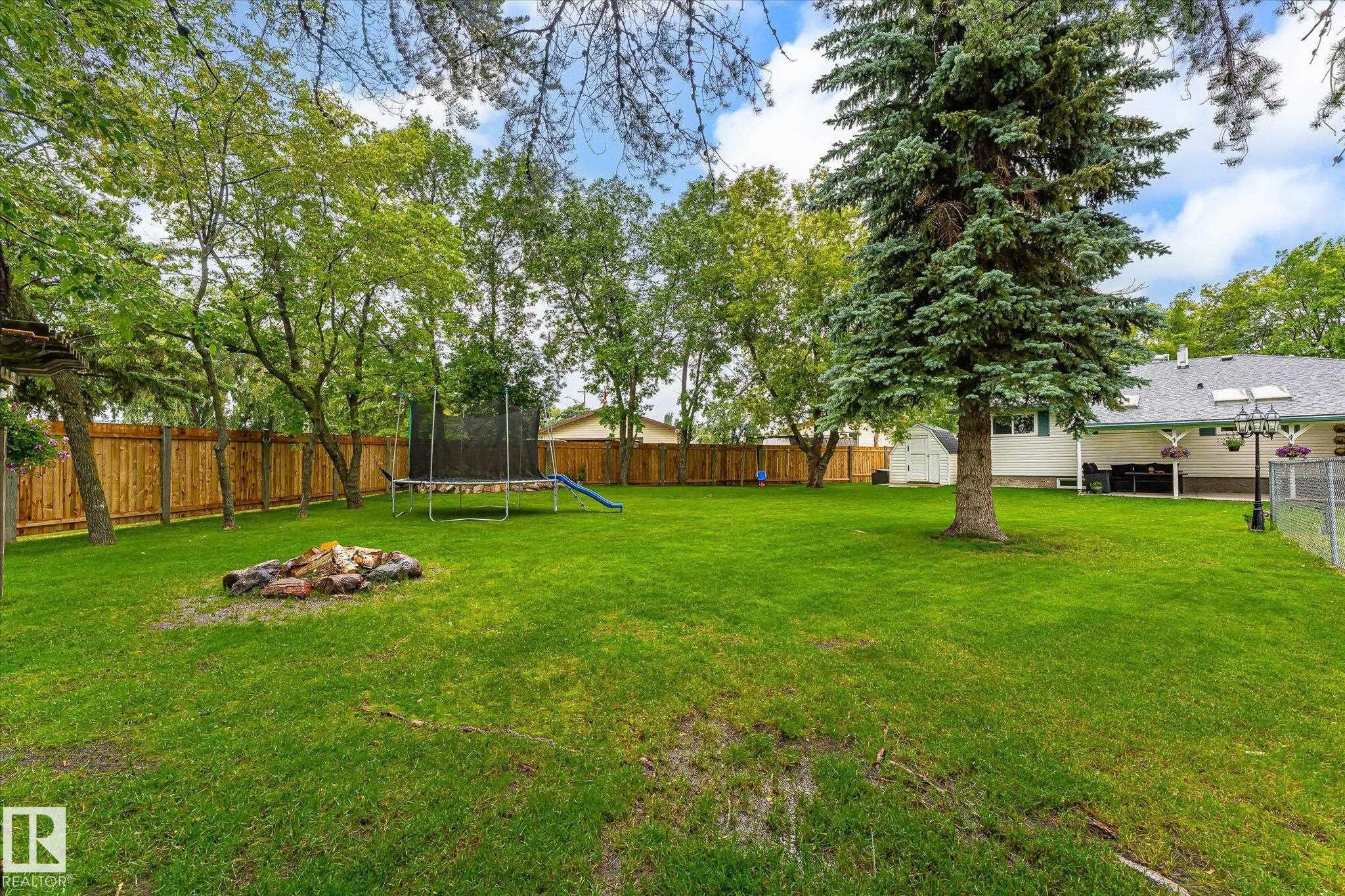 Expansive backyard featuring a lush lawn, a dedicated fire pit area, mature trees providing natural shade, a full wood privacy fence, and a white storage shed - 4921 47 Avenue, Legal, AB - Outdoor With Backyard