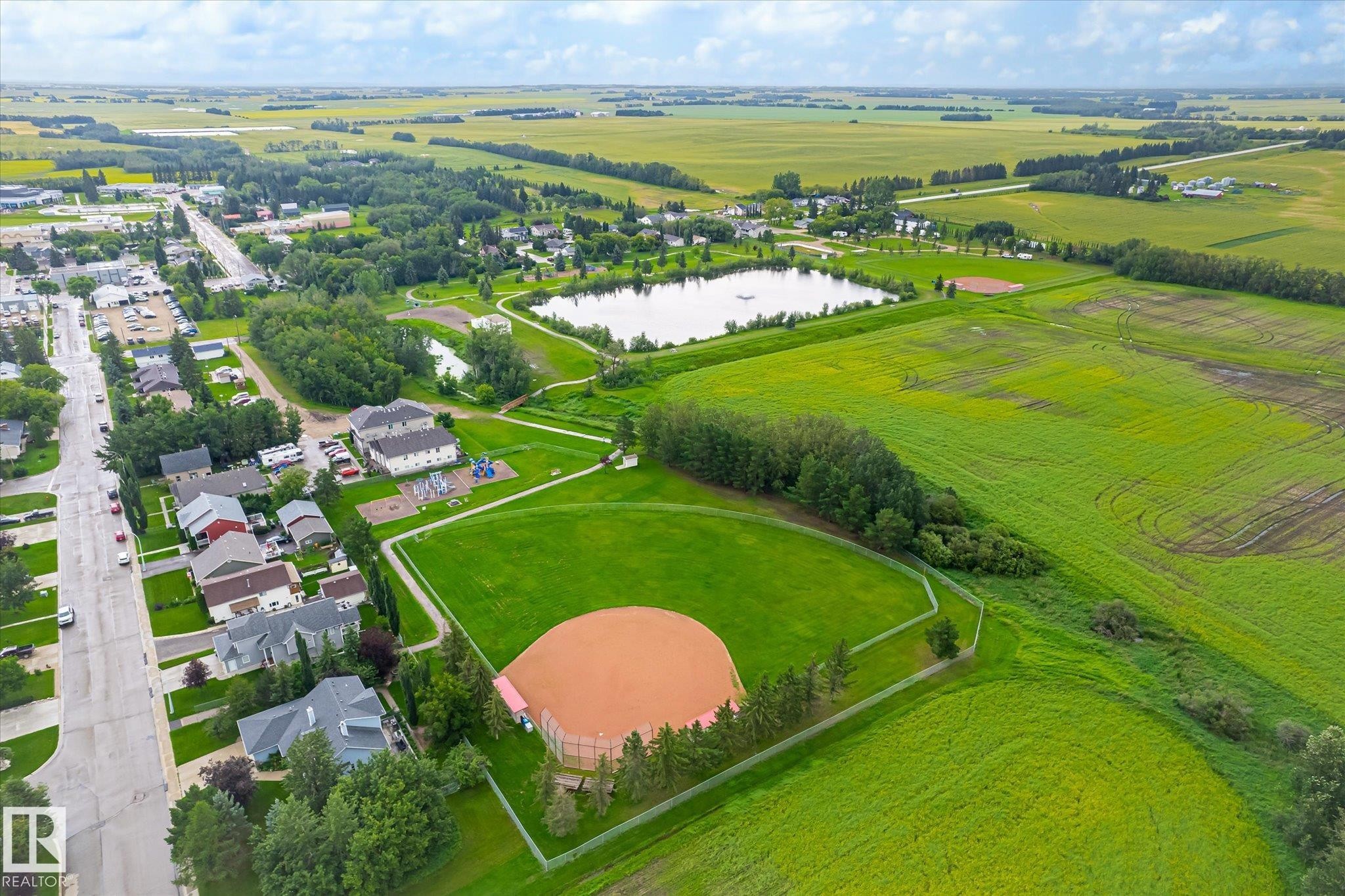 Aerial perspective showcasing a residential area with single-family homes, a tranquil pond, and verdant agricultural fields - 4921 47 Avenue, Legal, AB - Outdoor With View