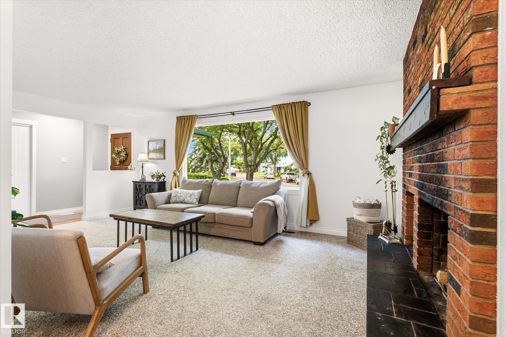 Spacious living area featuring a brick fireplace with a dark mantel, a large picture window, light-toned carpeting, and white walls - 4921 47 Avenue, Legal, AB - Indoor Photo Showing Living Room With Fireplace