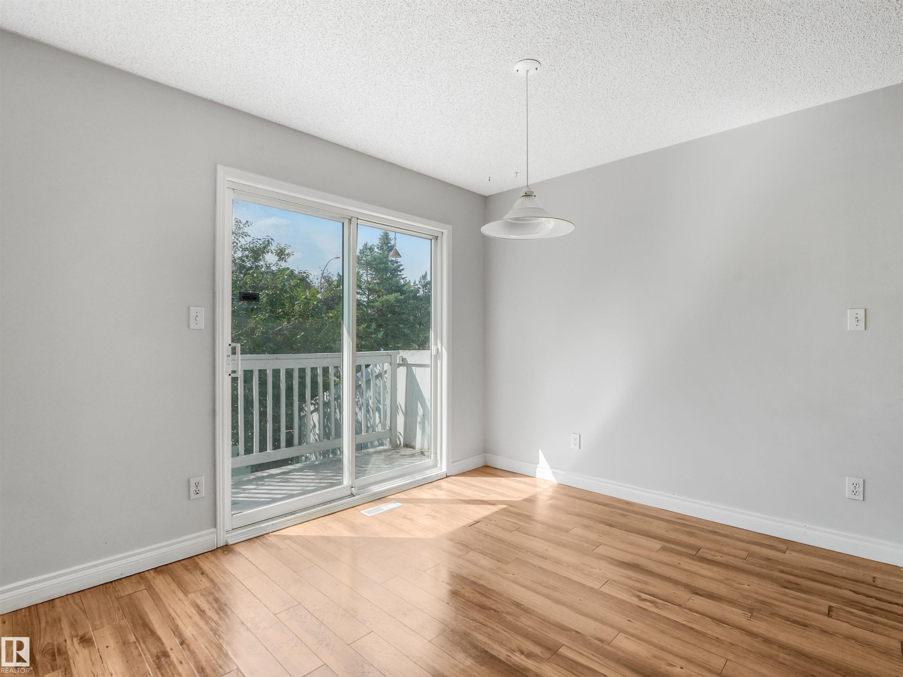 Sliding glass doors opening to a white-railed balcony, wood-finish flooring, a white pendant light fixture, and light gray walls - #2 3311 58 St Nw, Edmonton, AB - Indoor Photo Showing Other Room