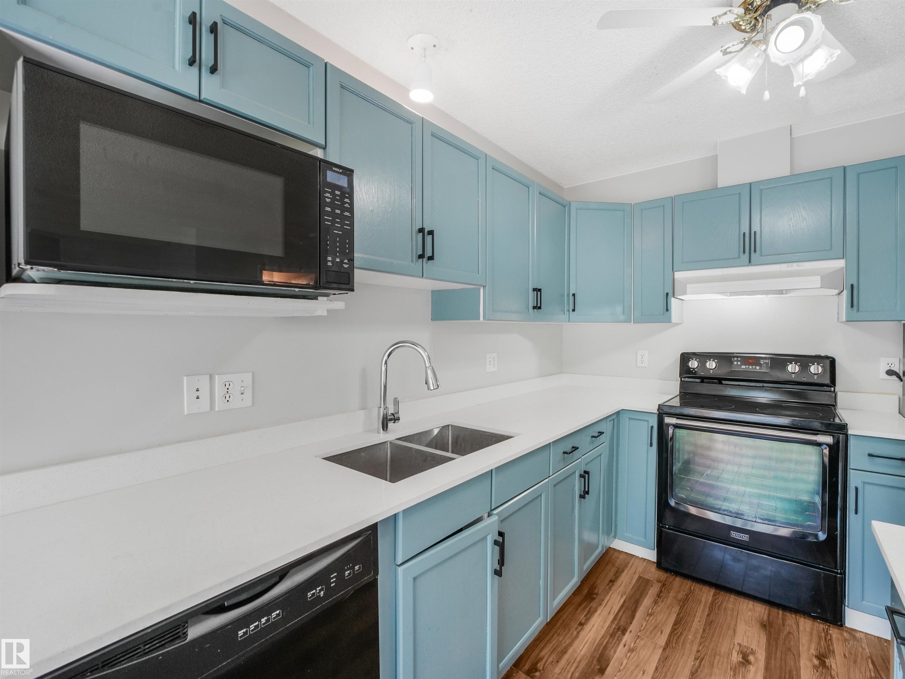 Kitchen featuring light blue cabinetry with dark hardware, white countertops, wood-finish flooring, a stainless steel double basin sink with a gooseneck faucet, and a black over-the-range microwave - #2 3311 58 St Nw, Edmonton, AB - Indoor Photo Showing Kitchen With Double Sink