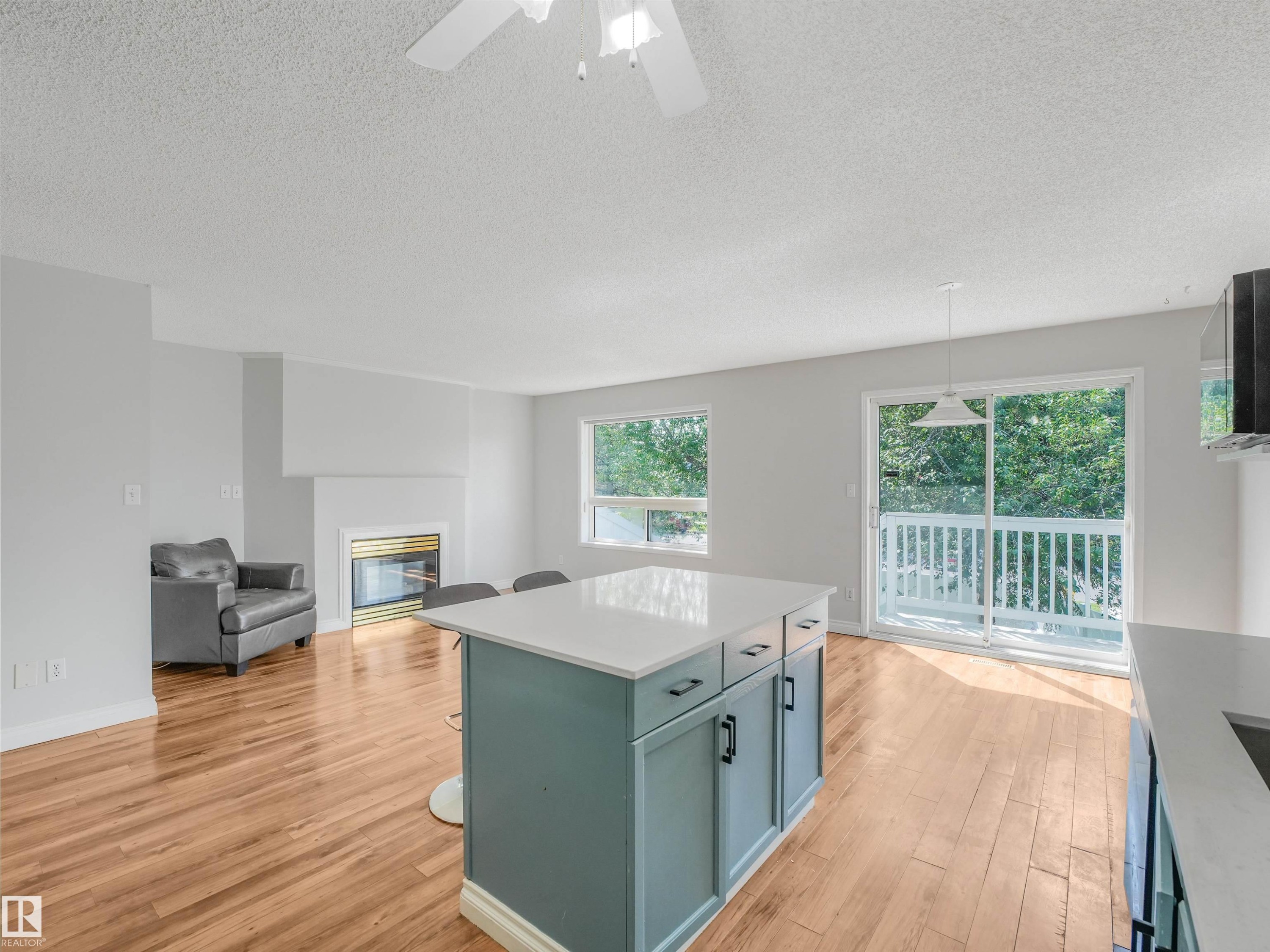 Open-concept living area featuring a fireplace, wood-finish flooring, a kitchen island with seating, a ceiling fan, and sliding glass doors leading to a private deck - #2 3311 58 St Nw, Edmonton, AB - Indoor Photo Showing Other Room With Fireplace