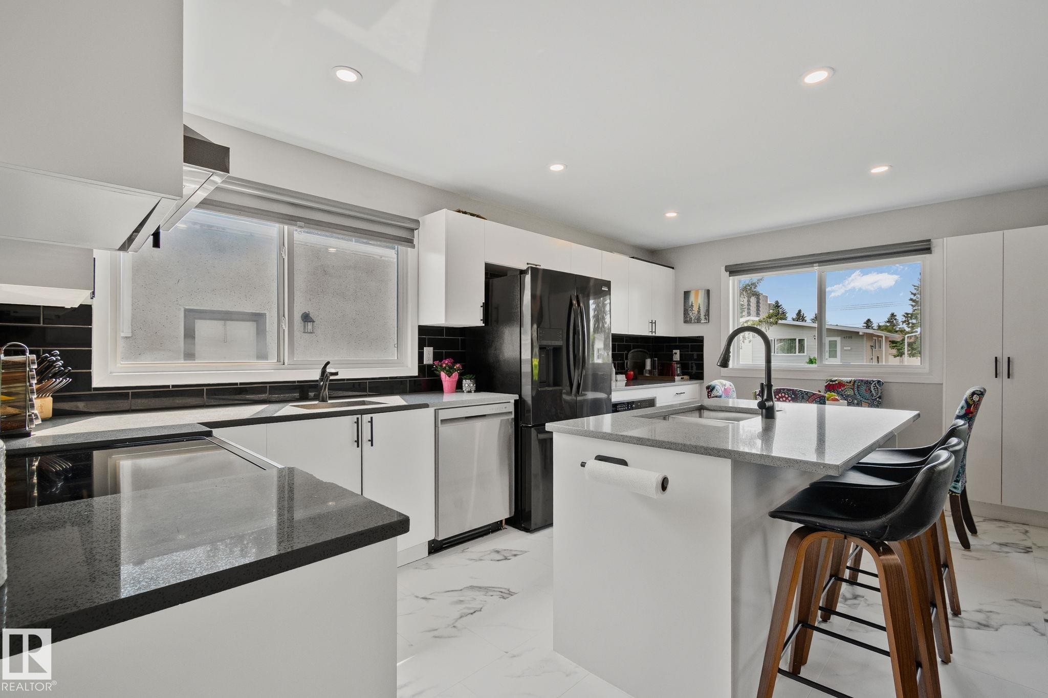 Modern kitchen featuring an island with seating, dark countertops, white cabinetry, a black tile backsplash, and stainless steel appliances - 4723 105 Street Nw, Edmonton, AB - Indoor Photo Showing Kitchen