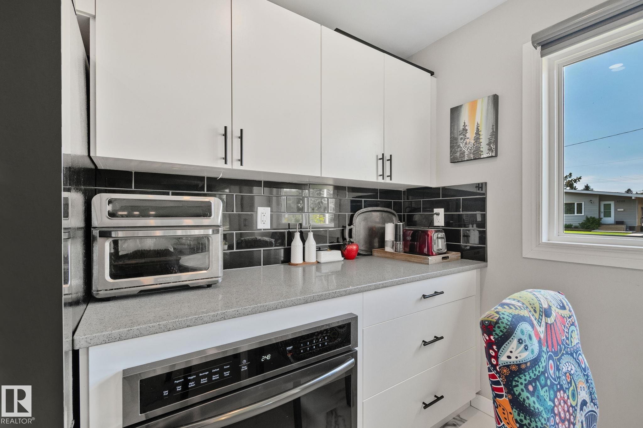 White upper and lower cabinetry with matte black hardware, black subway tile backsplash, light grey countertop, and a built-in stainless steel oven - 4723 105 Street Nw, Edmonton, AB - Indoor Photo Showing Kitchen