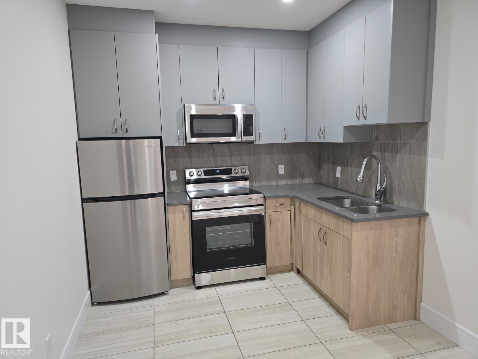 Kitchen featuring stainless steel appliances, grey upper cabinetry, wood-finish lower cabinetry, a double basin sink with gooseneck faucet, and a tiled backsplash - 228 36 Street Sw, Edmonton, AB - Indoor Photo Showing Kitchen With Double Sink