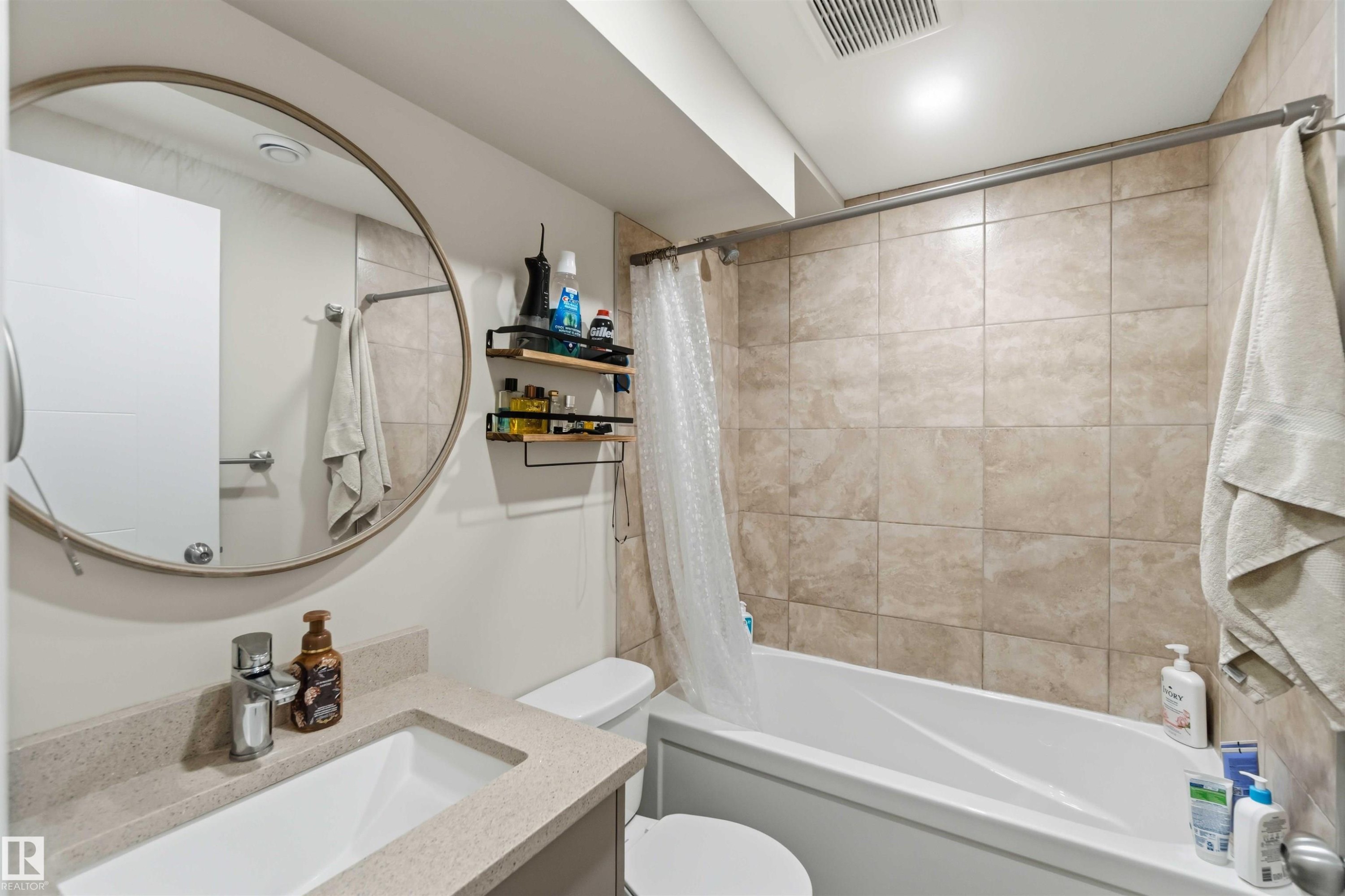 Bathroom featuring a white vanity with a rectangular undermount sink, a round wall-mounted mirror, a tiled shower-over-tub with a curtain rod, and light-toned wall tiling - 11351 35A Avenue Nw, Edmonton, AB - Indoor Photo Showing Bathroom