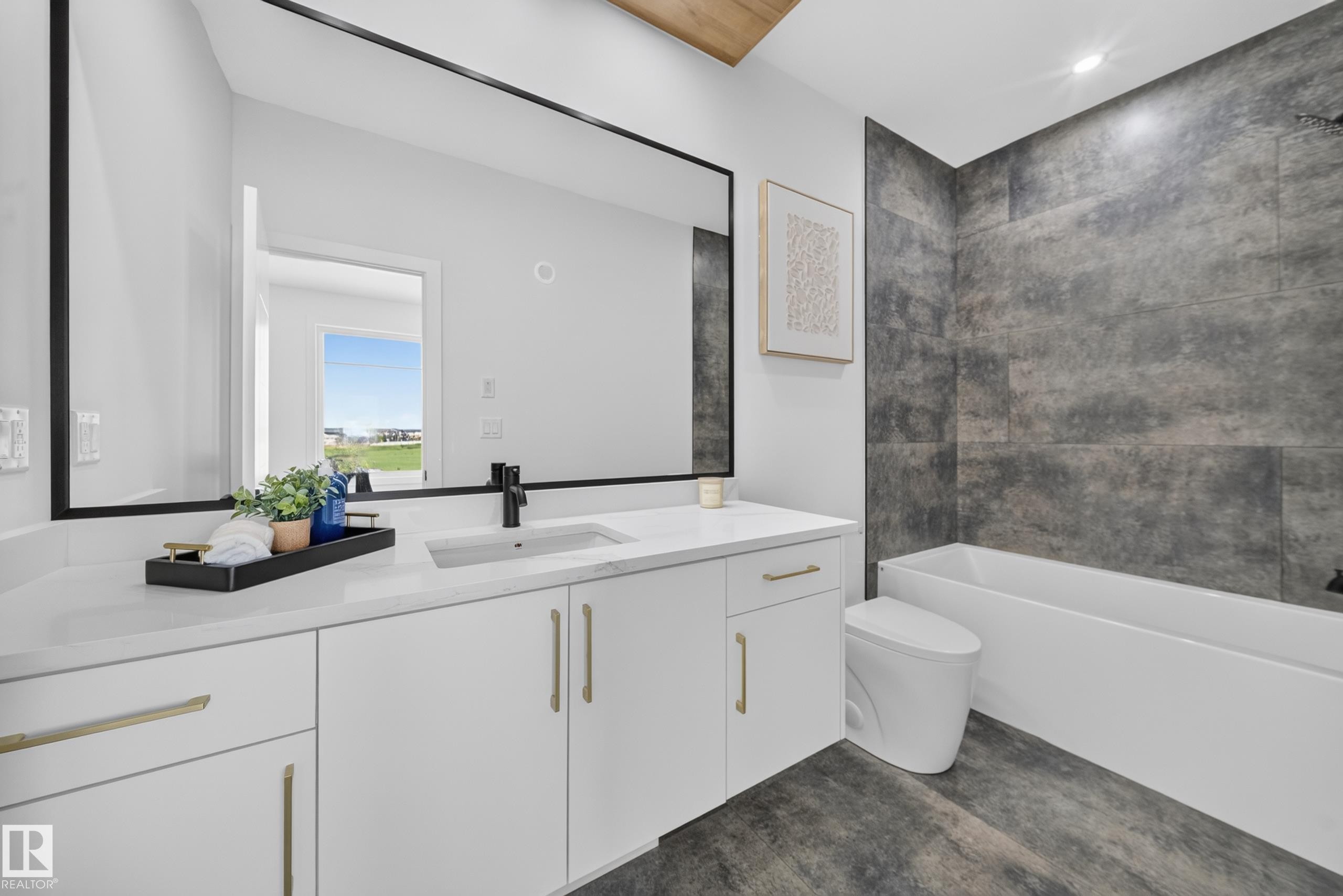Contemporary bathroom featuring an expansive vanity with a white countertop and gold-finish hardware, a large frameless mirror, a built-in tub with dark gray tiled surround, and wood-finish flooring - 423 Robinson Bv, Leduc, AB - Indoor Photo Showing Bathroom