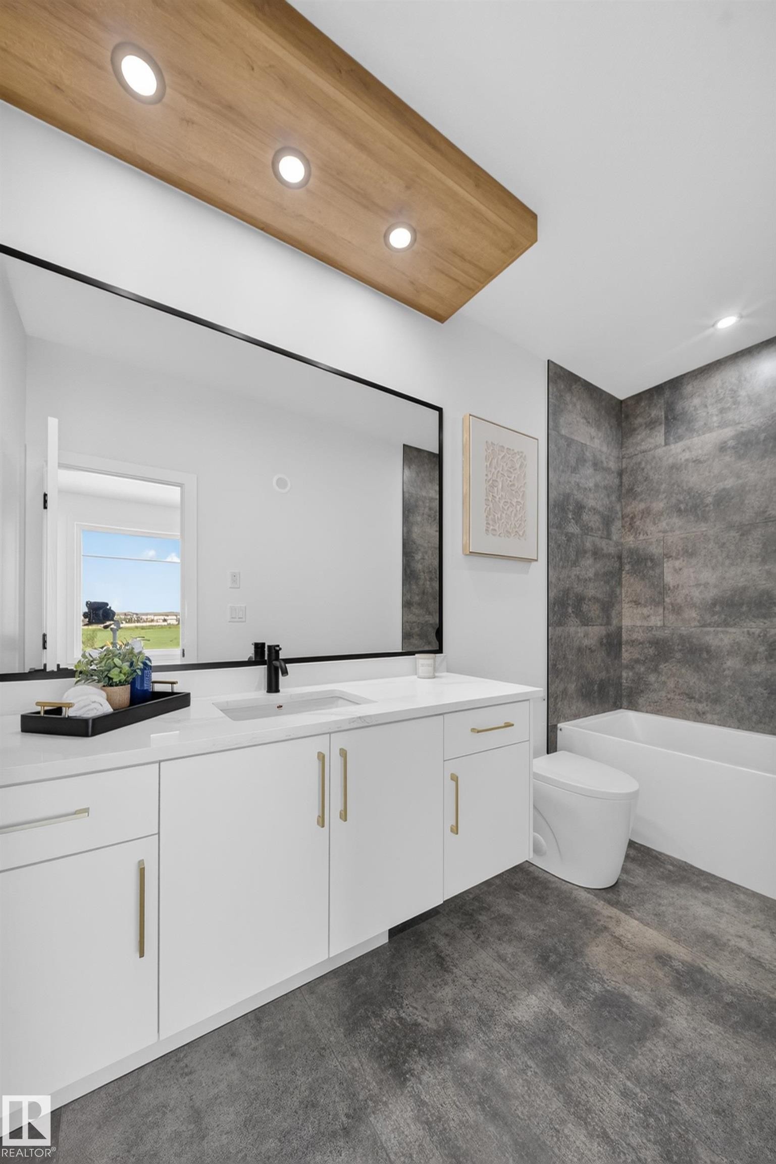 Modern bathroom featuring a double vanity with white cabinetry and gold-tone hardware, a large rectangular mirror with a black frame, a built-in tub with dark gray tile surround, and a wood-finish ceiling accent with recessed lighting - 423 Robinson Bv, Leduc, AB - Indoor Photo Showing Bathroom