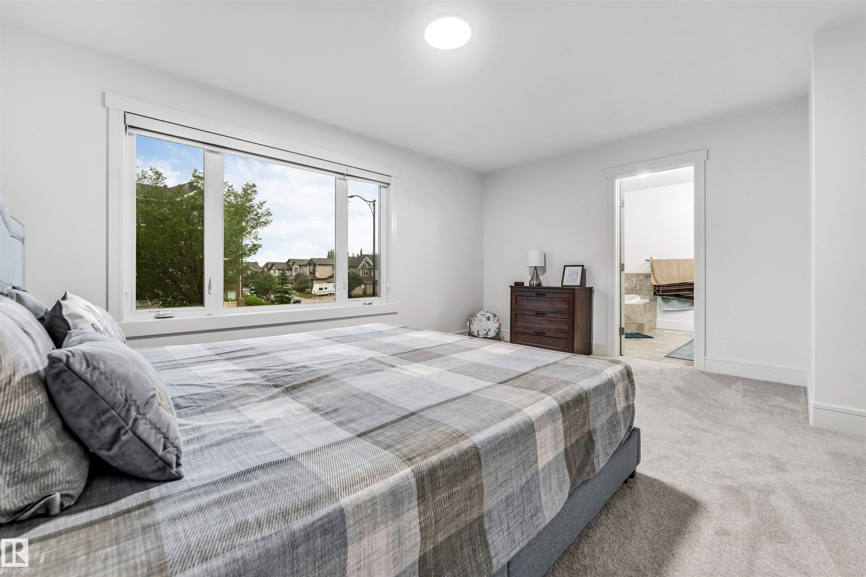 Expansive bedroom featuring light gray carpeting, a large picture window, and a white framed doorway opening to a bathroom with tiled flooring - 6727 20 Ave Sw, Edmonton, AB - Indoor Photo Showing Bedroom