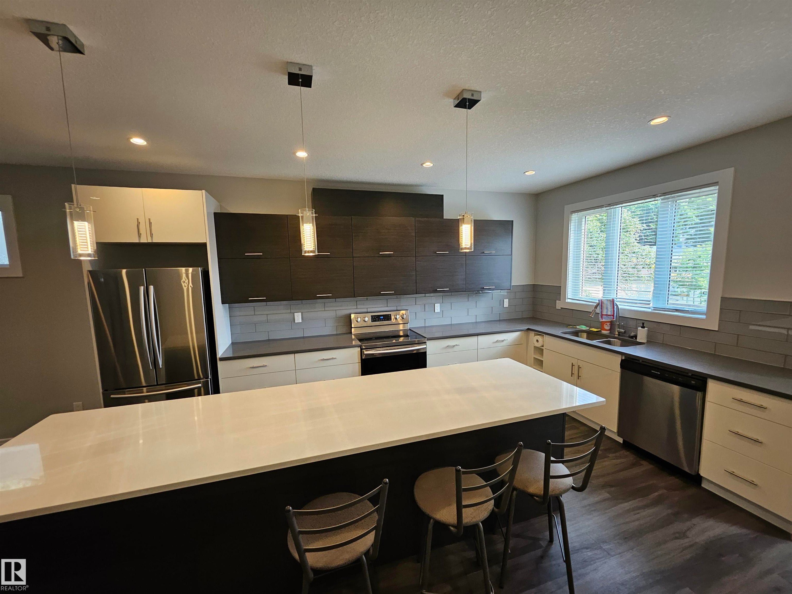 Contemporary kitchen featuring a large island with white countertop, dark wood-finish flooring, stainless steel appliances, and sleek pendant lighting - 9311 66 Avenue Nw, Edmonton, AB - Indoor Photo Showing Kitchen With Double Sink With Upgraded Kitchen