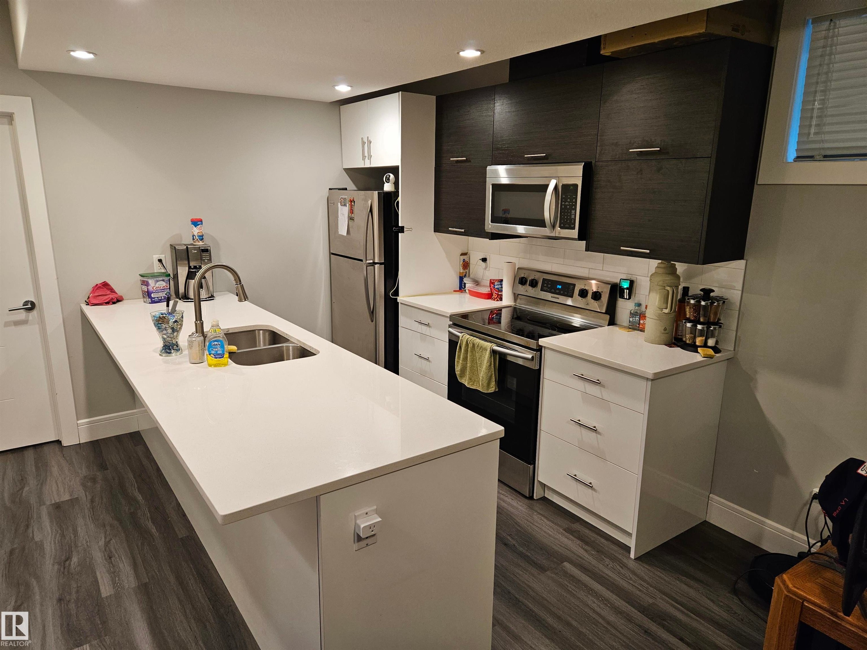 Contemporary kitchen featuring a large island with an undermount sink, stainless steel appliances, wood-finish flooring, recessed lighting, and mixed cabinetry with white uppers and dark lower cabinets - 9311 66 Avenue Nw, Edmonton, AB - Indoor Photo Showing Kitchen With Double Sink