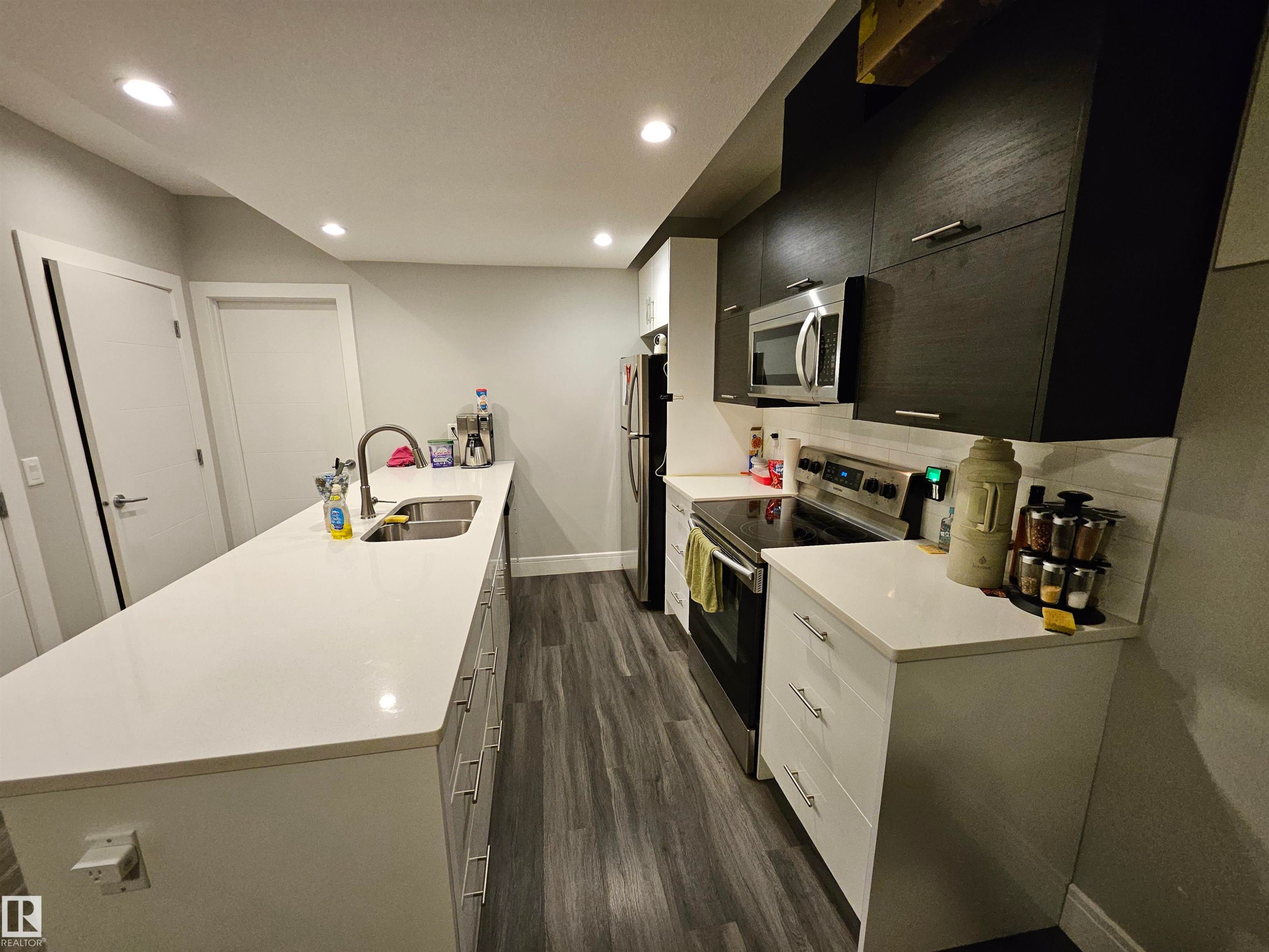 Contemporary kitchen featuring a spacious island with an undermount sink, white countertops, dark wood-finish upper cabinetry, stainless steel appliances, and wood-finish flooring - 9311 66 Avenue Nw, Edmonton, AB - Indoor Photo Showing Kitchen With Double Sink
