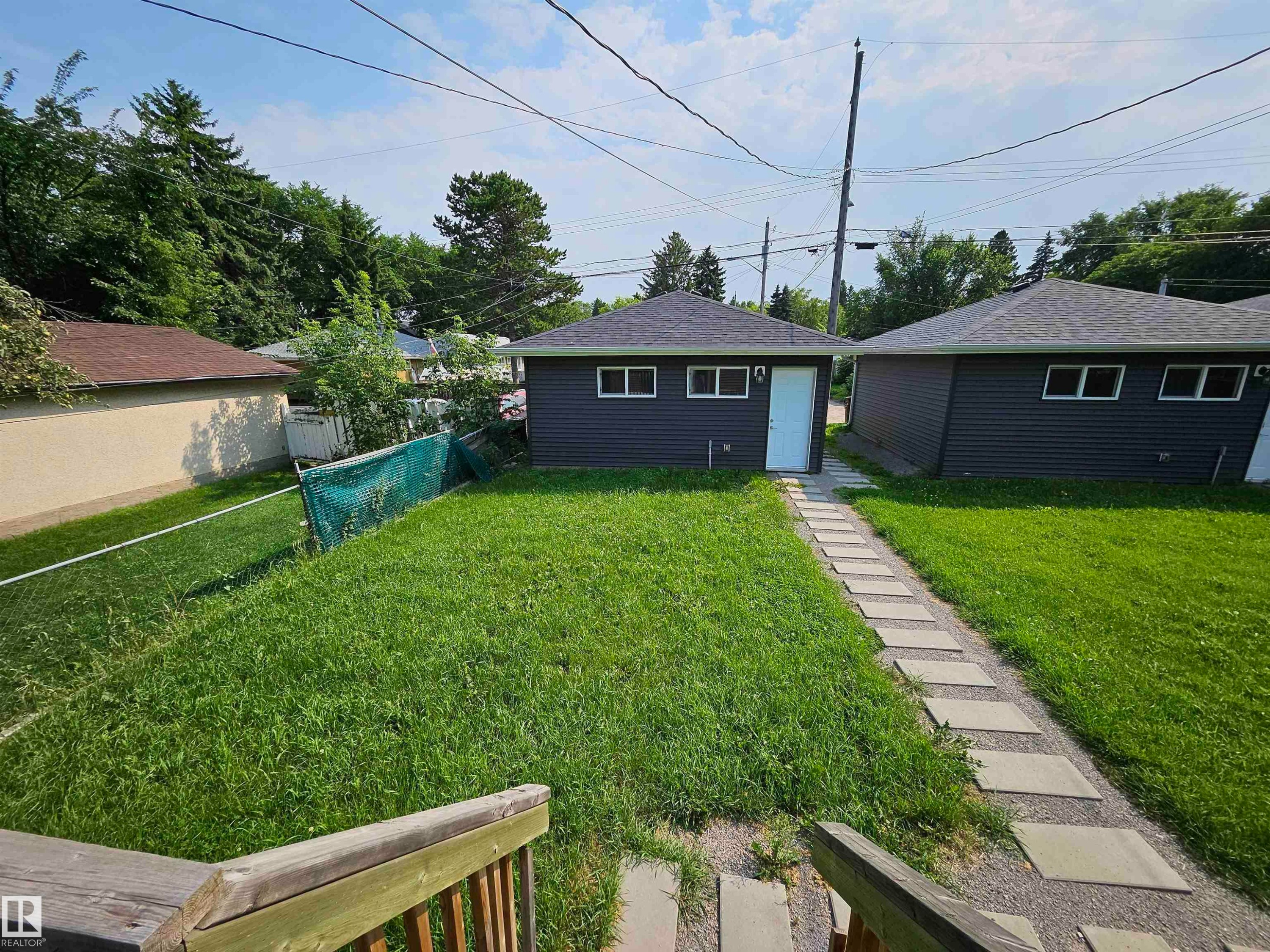 Expansive grassy yard area with a paver walkway leading to a detached structure featuring dark siding, a light-colored door, and multiple windows - 9311 66 Avenue Nw, Edmonton, AB - Outdoor
