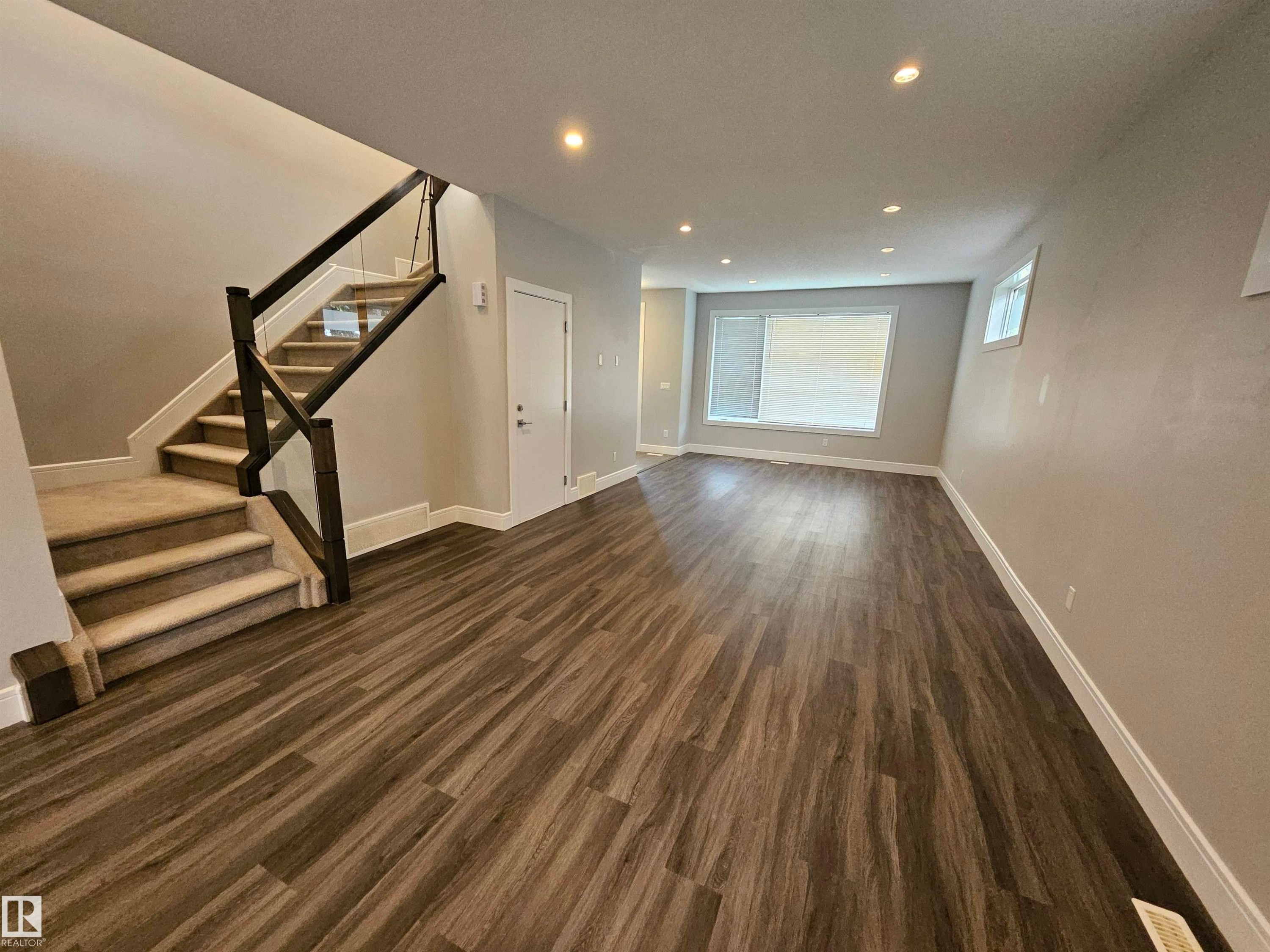 Spacious interior with wood-finish flooring, a contemporary staircase featuring glass paneling and dark wood railing, recessed ceiling lighting, and large windows - 9311 66 Avenue Nw, Edmonton, AB - Indoor Photo Showing Other Room