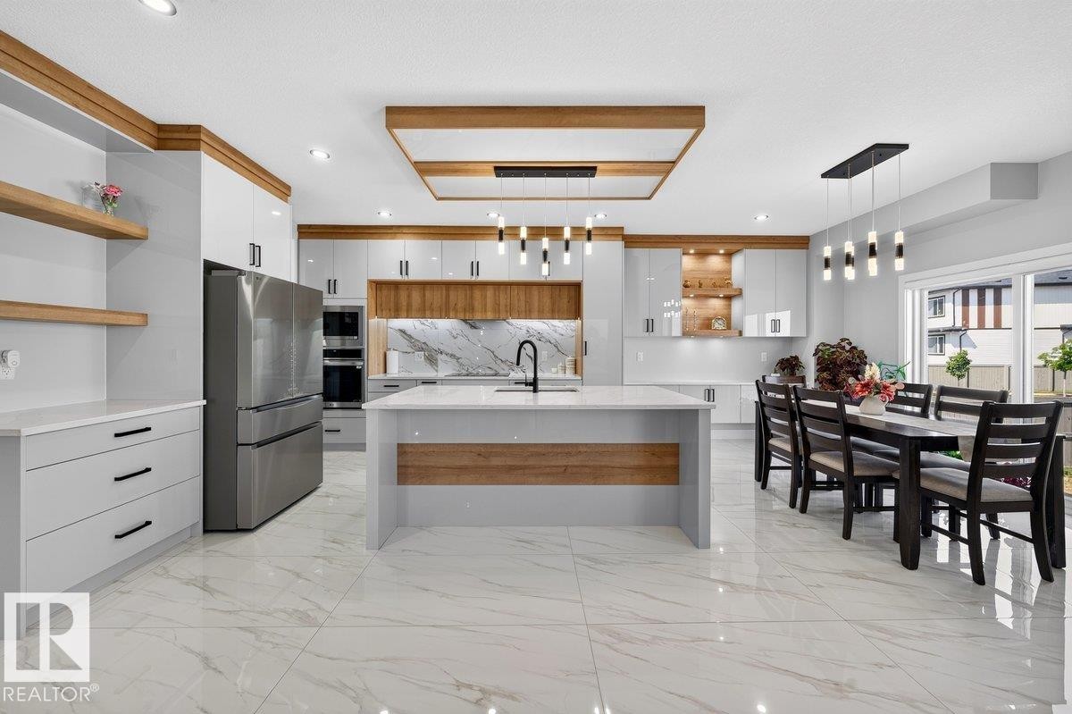 Open-concept kitchen and dining area featuring a central island, integrated stainless steel appliances, a marble-patterned backsplash, white cabinetry with wood-finish accents, and large format tile flooring - 6001 19 Street, Nisku, AB - Indoor