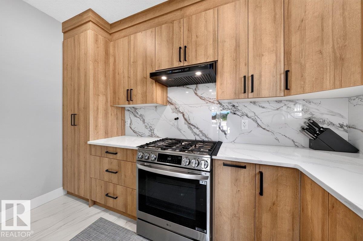 Modern kitchen featuring wood-finish cabinetry with black hardware, white countertops, a gas range with a black vent hood, and a white marble-patterned backsplash - 6001 19 Street, Nisku, AB - Indoor Photo Showing Kitchen