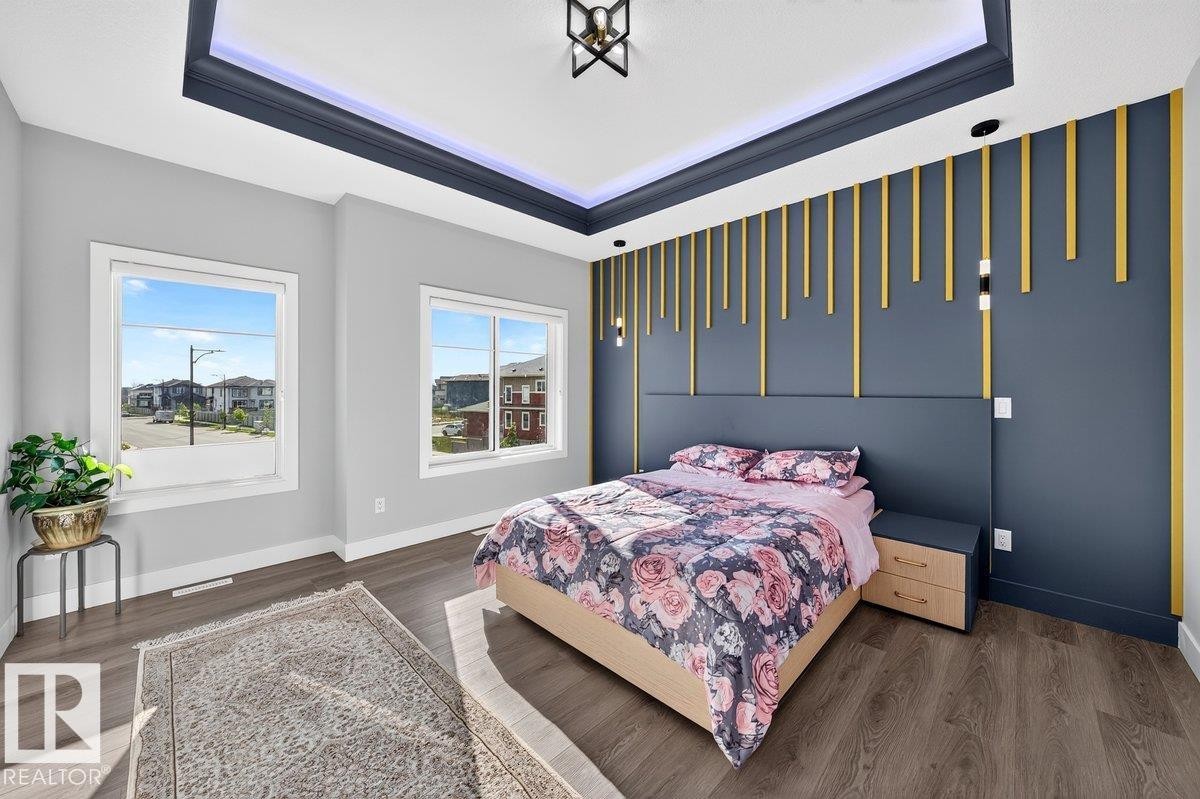 Spacious room featuring wood-finish flooring, two large windows, a tray ceiling with integrated lighting, and a decorative accent wall with vertical gold elements - 6001 19 Street, Nisku, AB - Indoor Photo Showing Bedroom