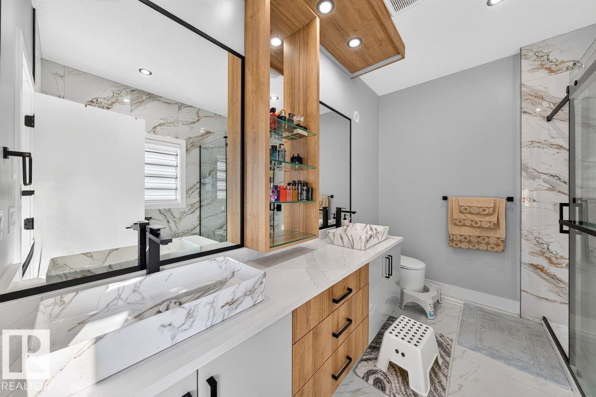 Bathroom featuring dual vessel sinks with matte black faucets, light wood-finish cabinetry, a large frameless mirror, recessed lighting, and a glass-enclosed shower with white and gold veined tile - 6001 19 Street, Nisku, AB - Indoor Photo Showing Bathroom