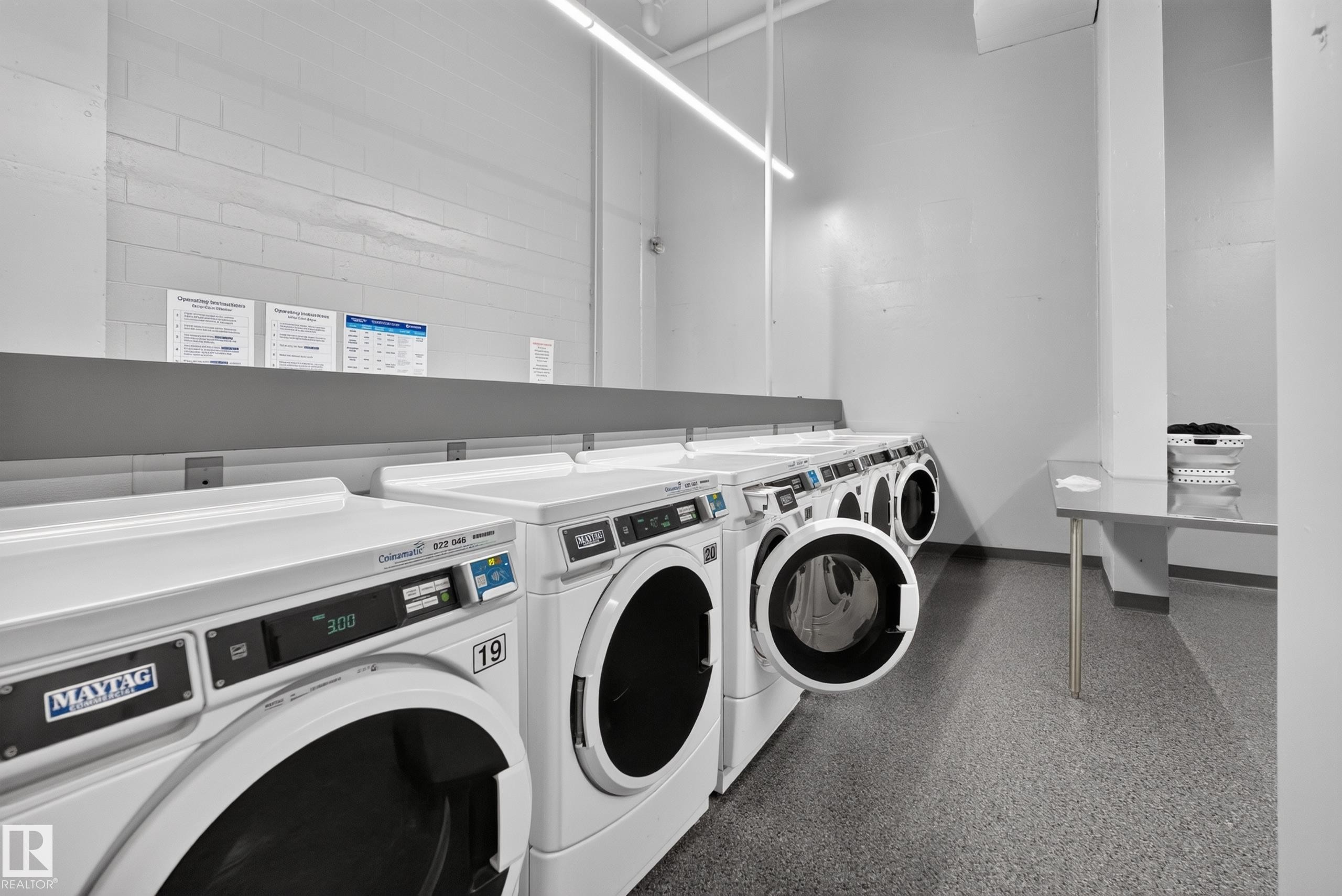 Laundry facility featuring multiple front-loading washing machines, durable flooring, and a folding counter - #1508 10883 Saskatchewan Dr Nw Nw, Edmonton, AB - Indoor Photo Showing Laundry Room