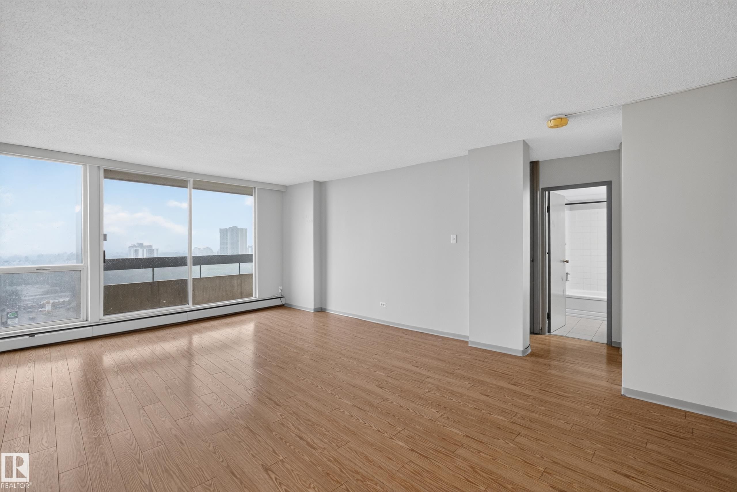 Expansive living area featuring large windows, wood-finish flooring, light gray wall paint, a built-in baseboard heating unit, and an open doorway to an interior space with tile flooring - #1508 10883 Saskatchewan Dr Nw Nw, Edmonton, AB - Indoor Photo Showing Other Room