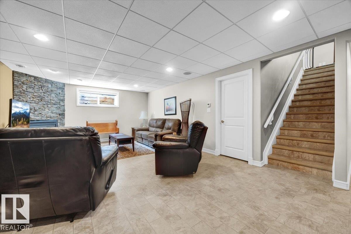 Finished lower level featuring light-toned flooring, a stone-accented wall with fireplace, recessed lighting, drop ceiling, and a staircase with a white railing - 928 Thompson Place Nw, Edmonton, AB - Indoor