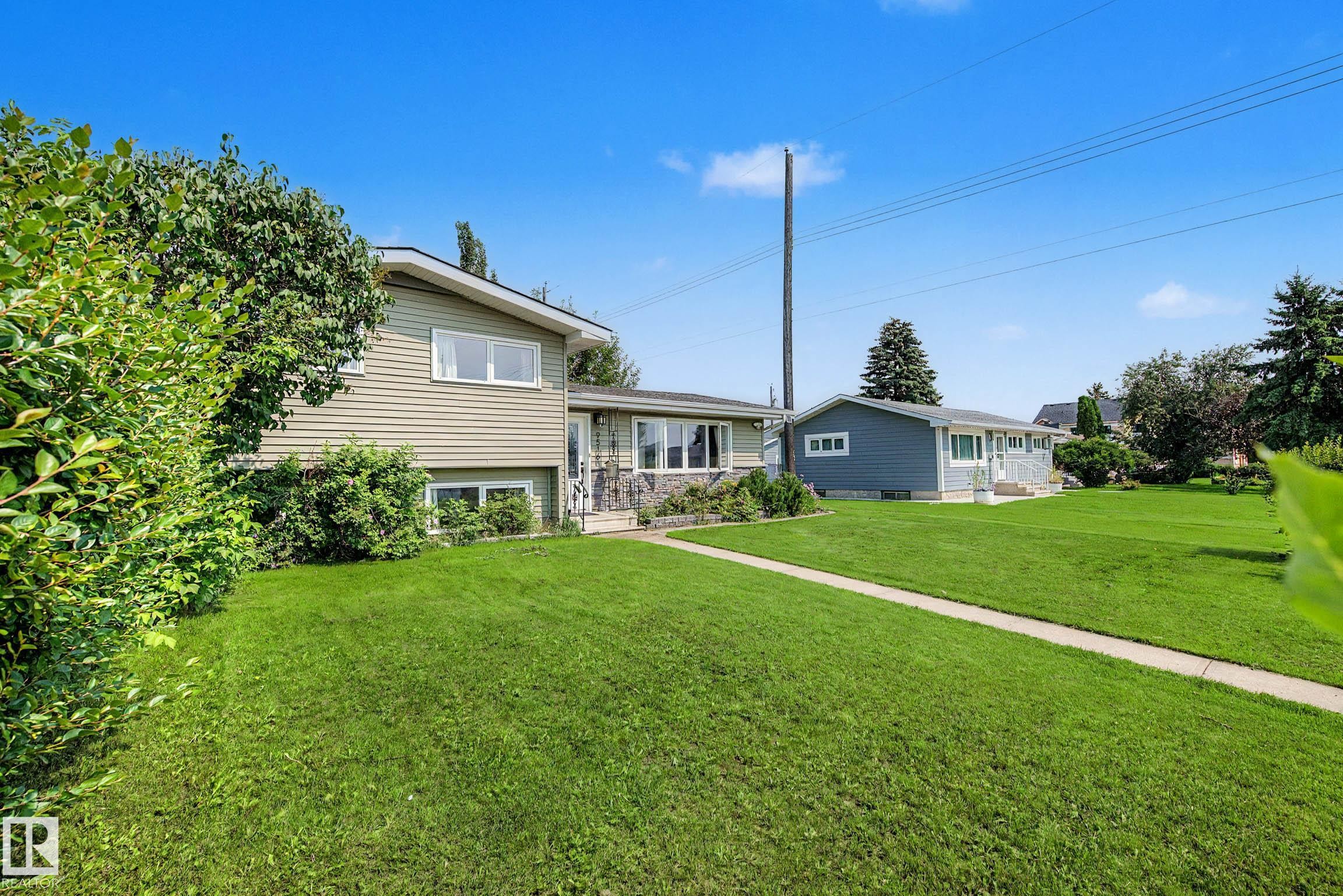 Expansive green lawn with a concrete pathway leading to a split-level residence featuring light-colored siding and a stone facade accent - 9516 129A Avenue Nw, Edmonton, AB