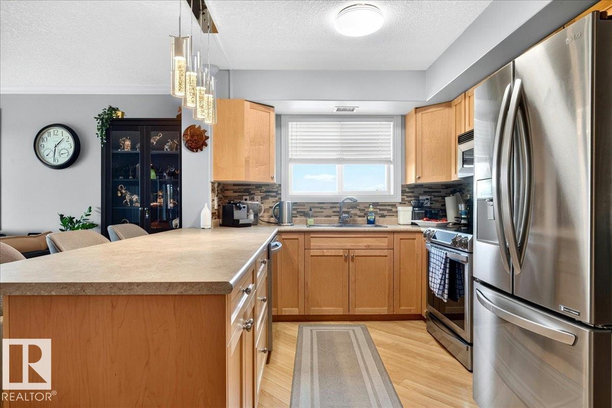 Kitchen featuring wood cabinetry, stainless steel appliances, an island with countertop seating, a mosaic tile backsplash, and wood-finish flooring - #306 13111 140 Av Nw, Edmonton, AB - Indoor Photo Showing Kitchen