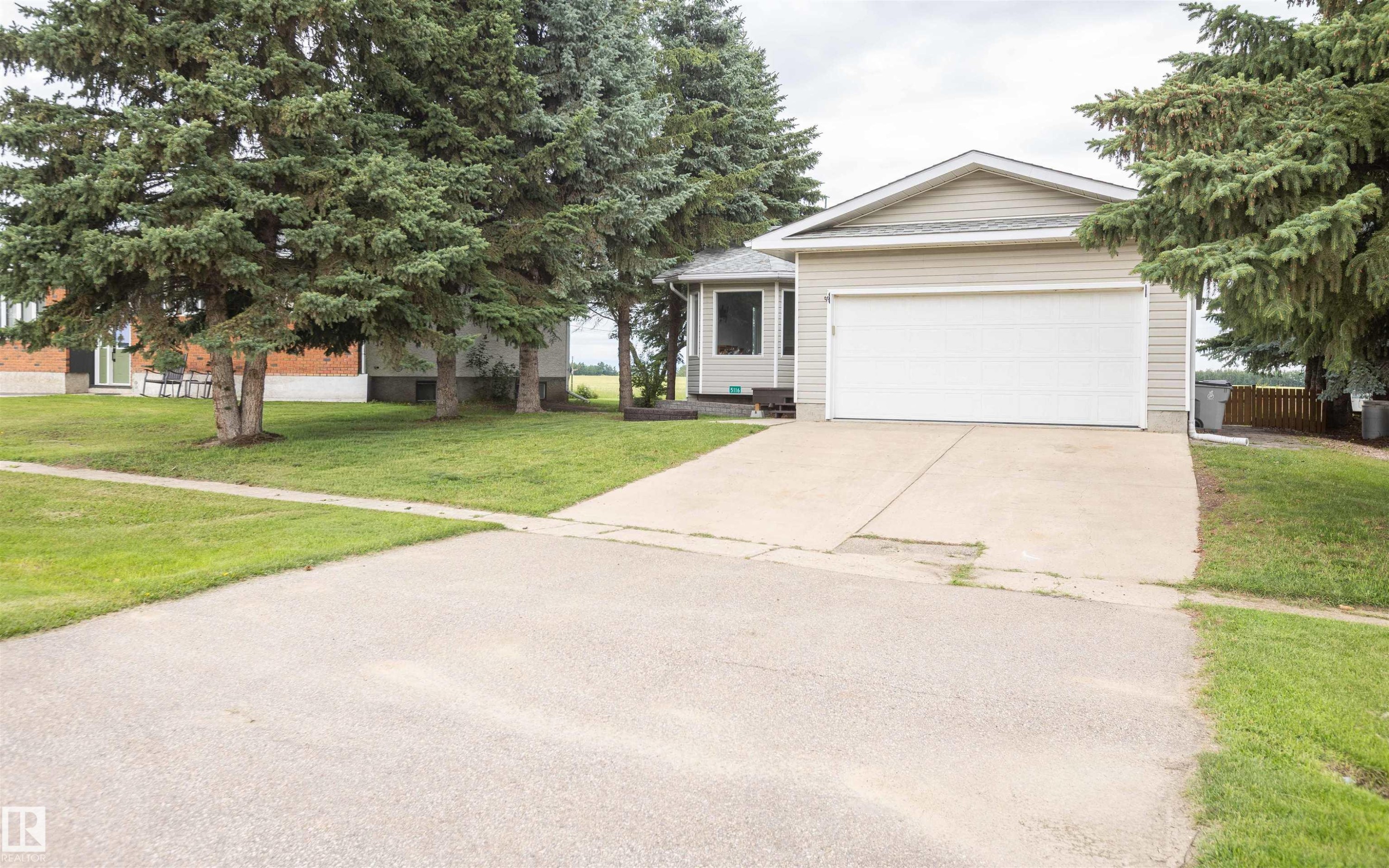 Exterior featuring a two-car garage with a white door, light-toned siding, a concrete driveway, and mature evergreen trees - 5116 51 Avenue, Pickardville, AB - Outdoor