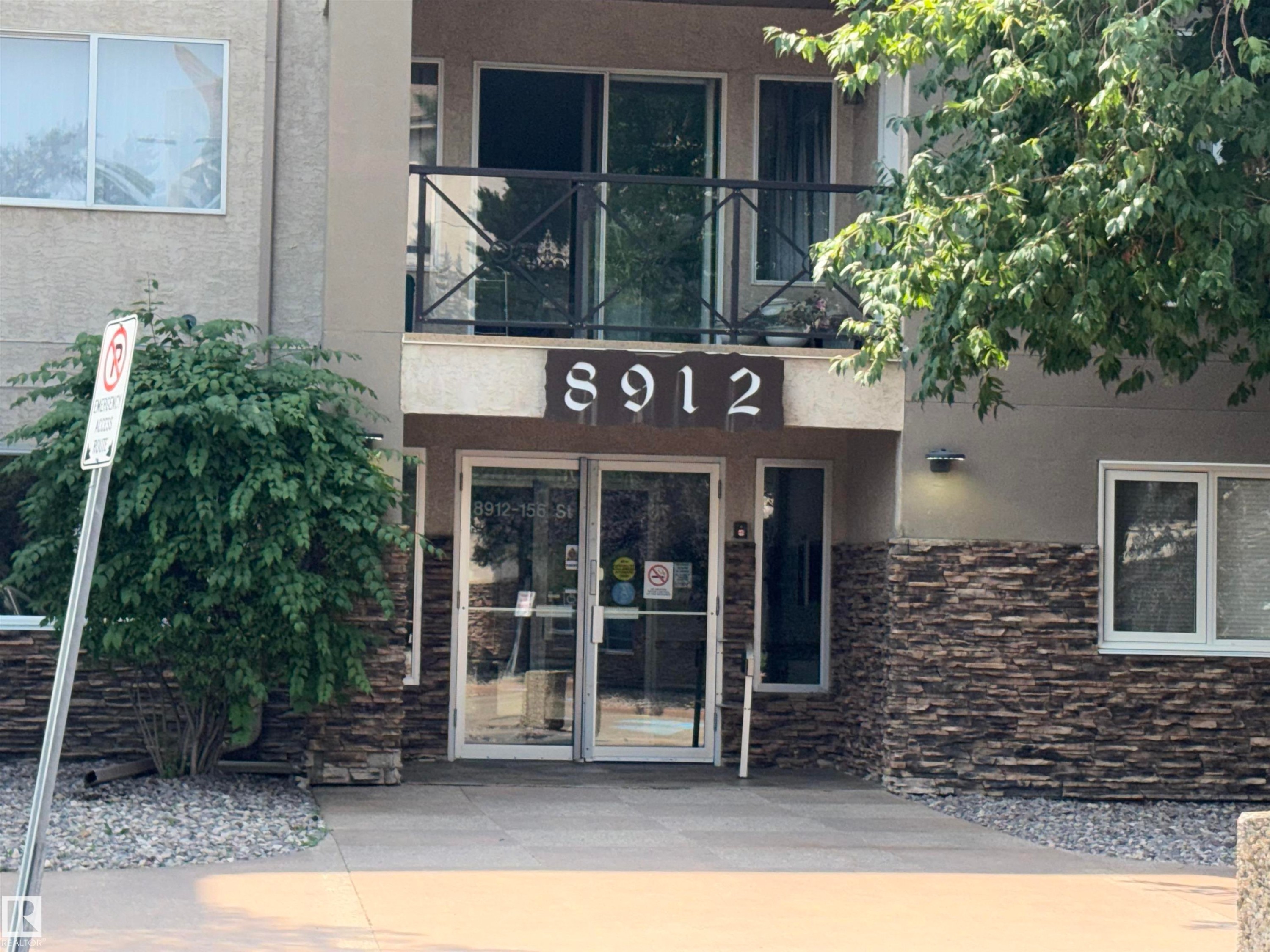 Building exterior featuring a stone facade, glass entry doors, and a second-story balcony with metal railings - #209 8912 156 St Nw, Edmonton, AB - Outdoor