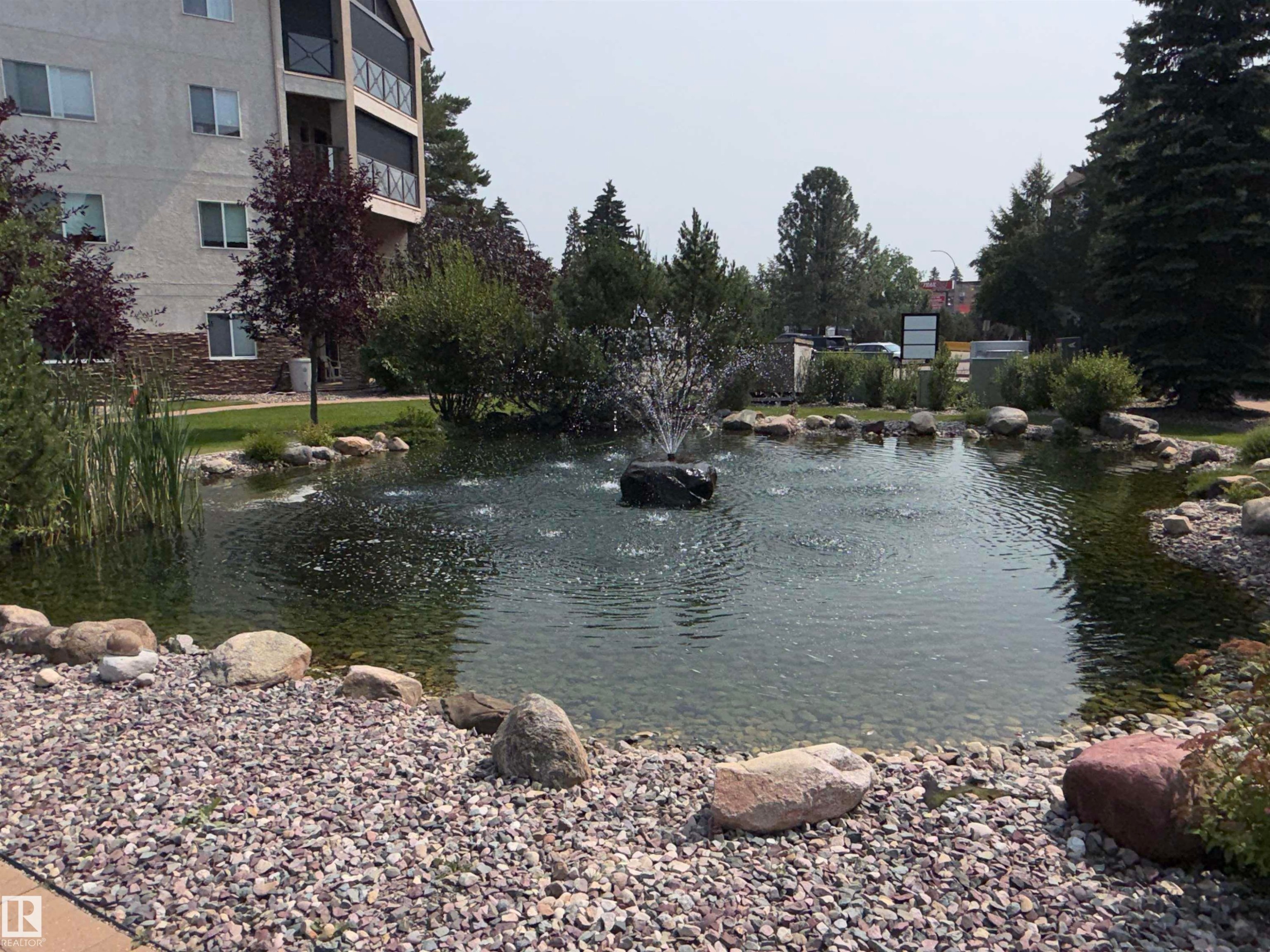 Landscaped pond with a central fountain feature, surrounded by decorative rocks and gravel - #209 8912 156 St Nw, Edmonton, AB - Outdoor With Body Of Water