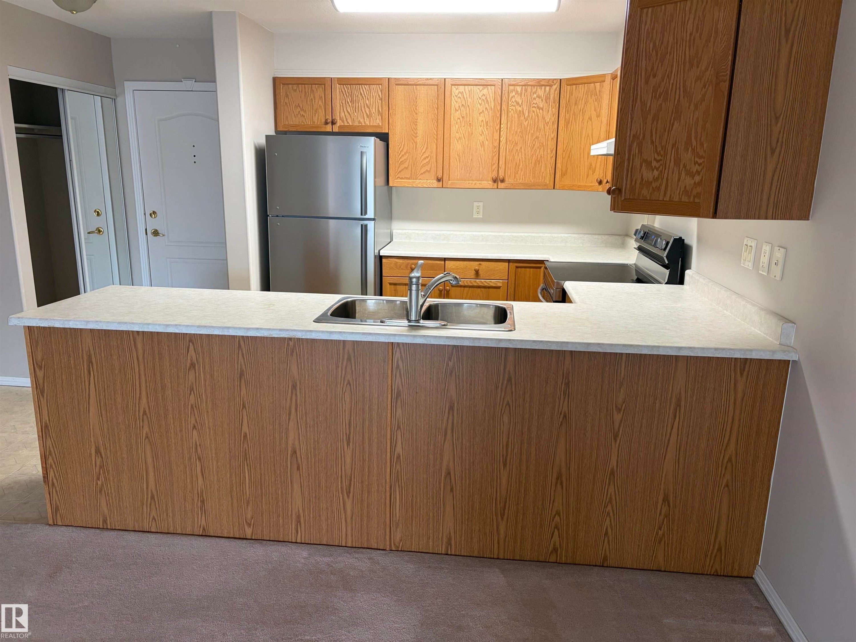 Kitchen featuring wood-finish cabinetry, stainless steel sink, light-toned countertops, and a full-size stainless steel refrigerator - #209 8912 156 St Nw, Edmonton, AB - Indoor Photo Showing Kitchen With Double Sink