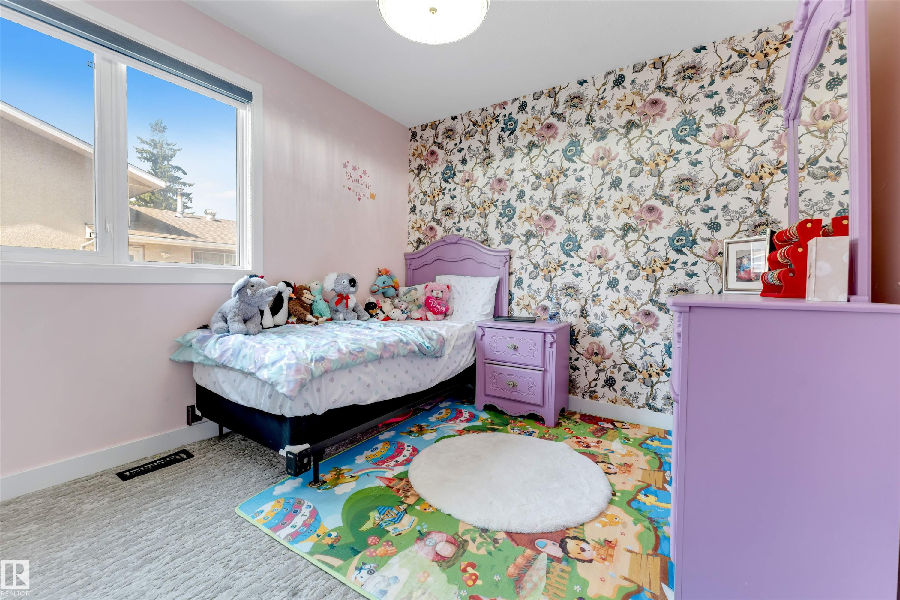 Light-filled bedroom featuring a large window, a floral accent wall, painted lavender furnishings, and soft carpet flooring - 7419 33 Avenue Nw, Edmonton, AB - Indoor Photo Showing Bedroom