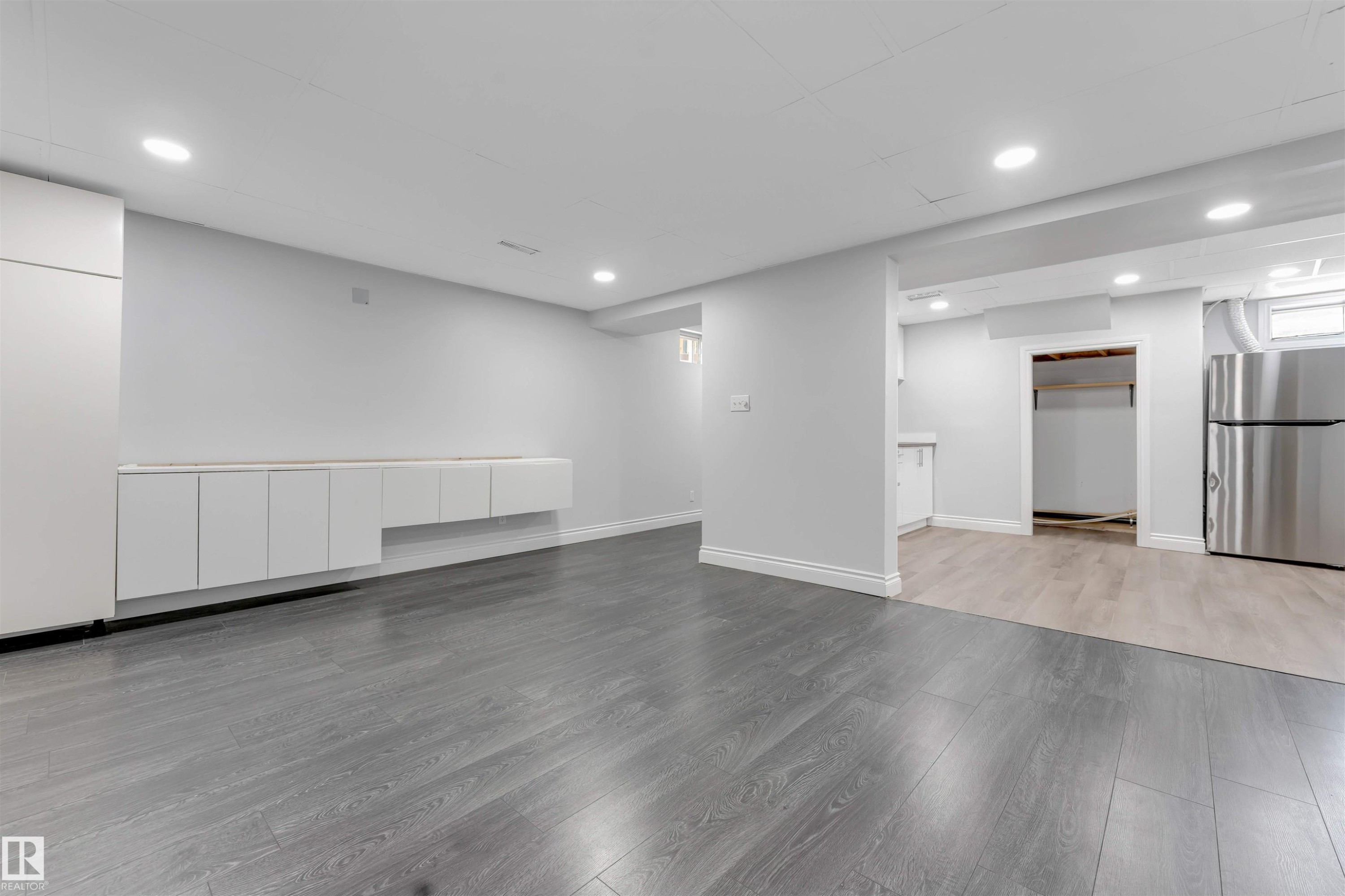 Spacious room with gray wood-finish flooring, white baseboards, recessed lighting, and a long built-in white cabinet unit - 7419 33 Avenue Nw, Edmonton, AB - Indoor
