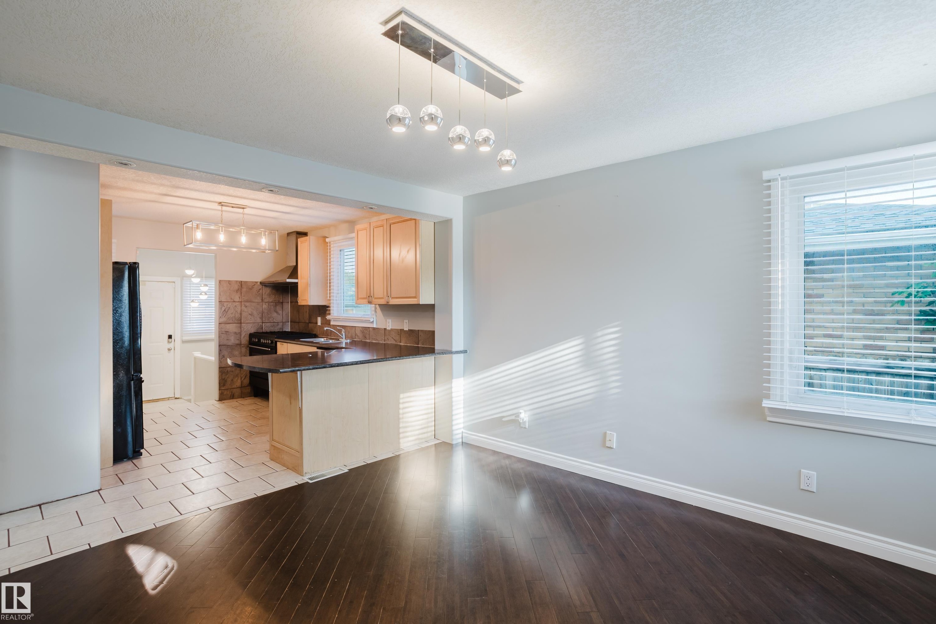 Open-concept living area featuring dark wood-finish flooring, a contemporary linear chandelier, and a bright window with blinds - 12431 81 Street Nw, Edmonton, AB - Indoor Photo Showing Kitchen