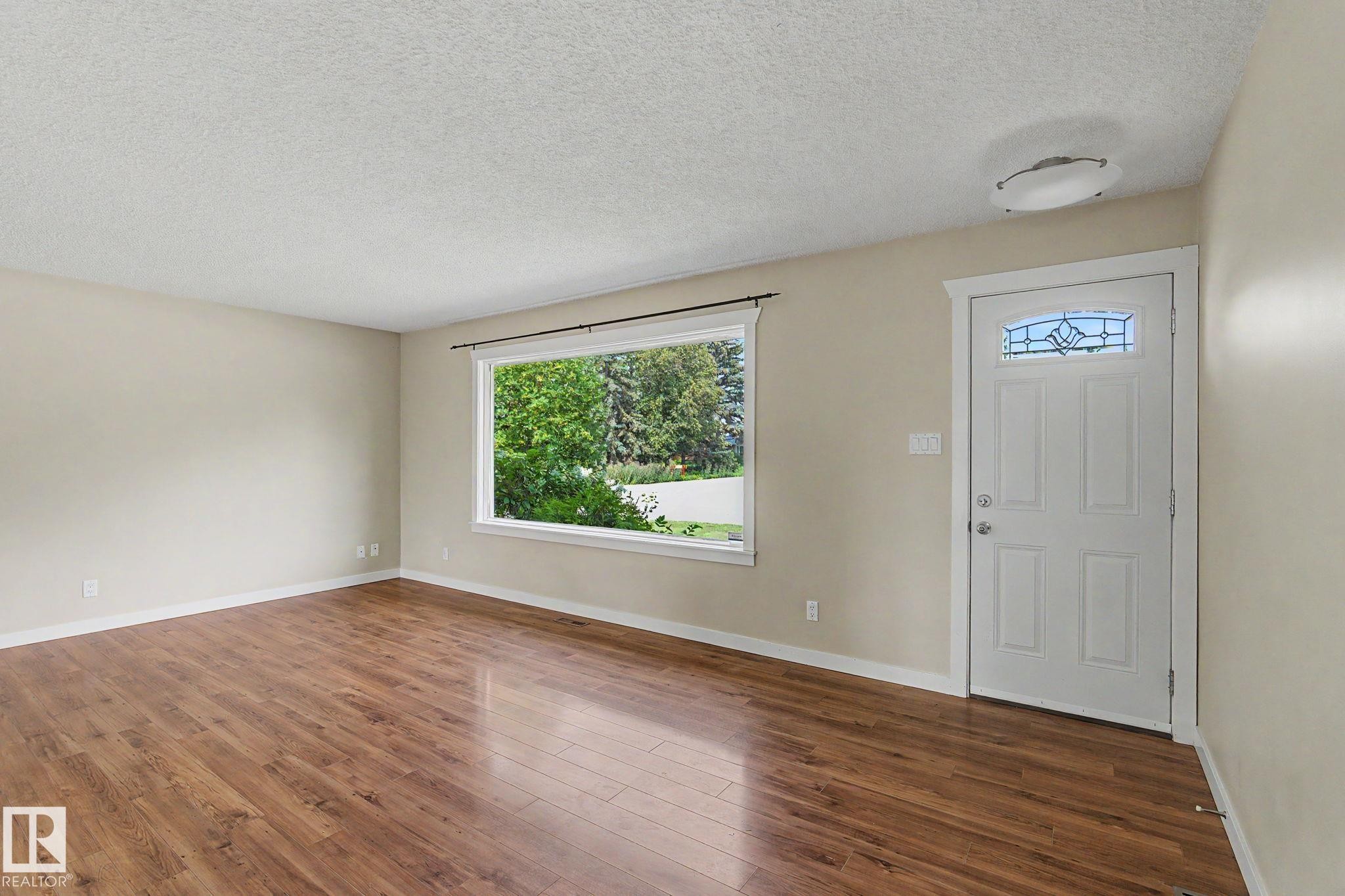Living area featuring wood-finish flooring, neutral wall tones, and a large front-facing window - 8823 130A Avenue Nw, Edmonton, AB - Indoor Photo Showing Other Room