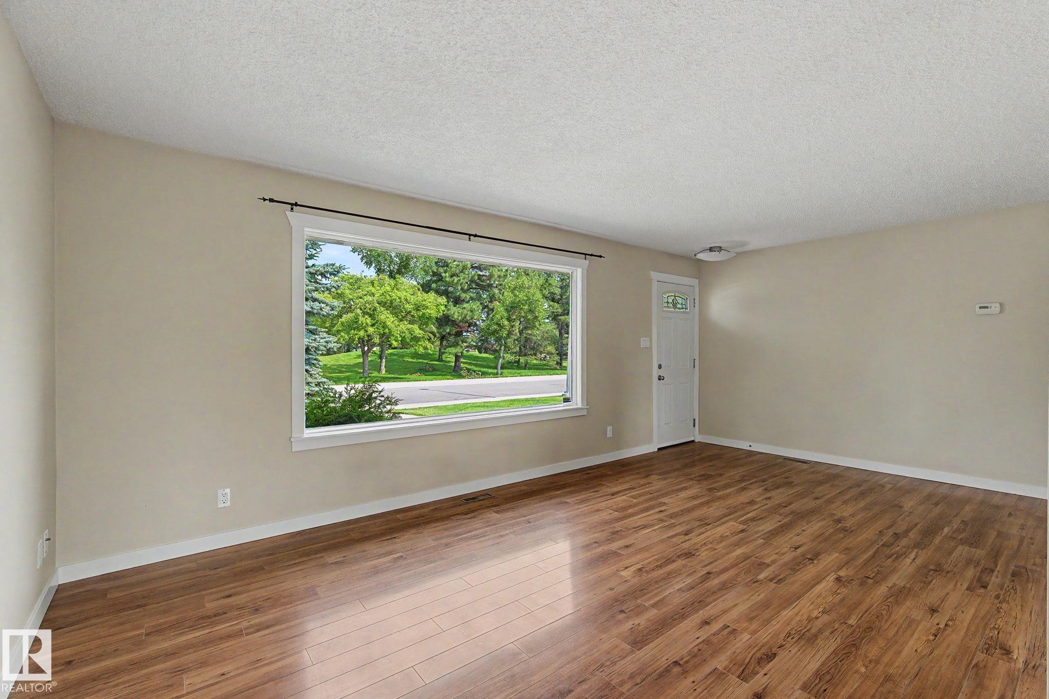 Bright interior space featuring a large picture window, wood-finish flooring, light neutral wall paint, white trim, and a white entry door with decorative glass - 8823 130A Avenue Nw, Edmonton, AB - Indoor Photo Showing Other Room