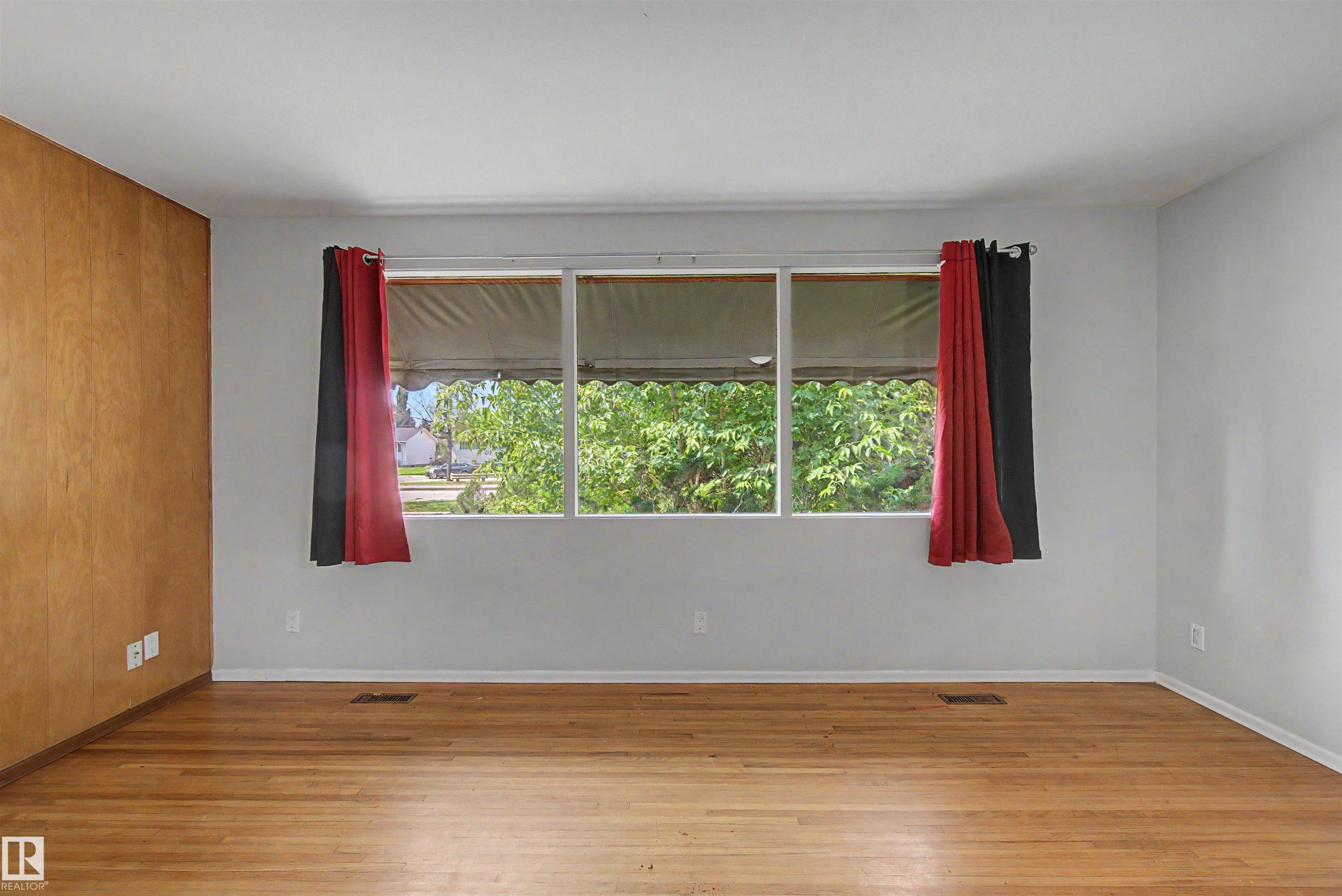 Spacious interior featuring a large multi-pane window, wood-finish flooring, a wood-paneled accent wall, and light gray painted walls - 8215 149 Street Nw, Edmonton, AB - Indoor Photo Showing Other Room