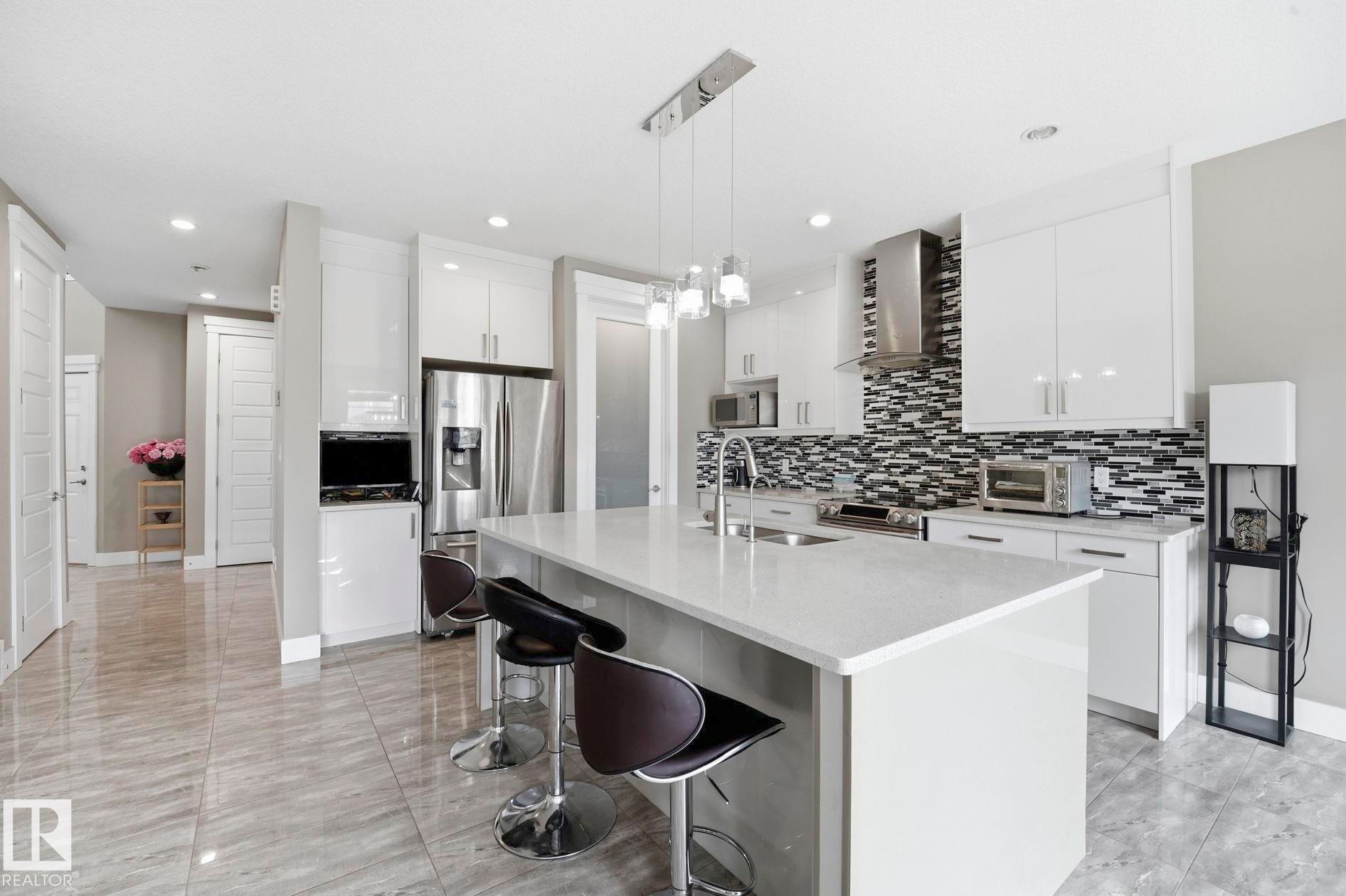 Modern kitchen featuring a large center island with integrated sink, stainless steel appliances, white cabinetry, and a monochrome mosaic tile backsplash - 1051 174 Street Sw, Edmonton, AB - Indoor Photo Showing Kitchen With Upgraded Kitchen