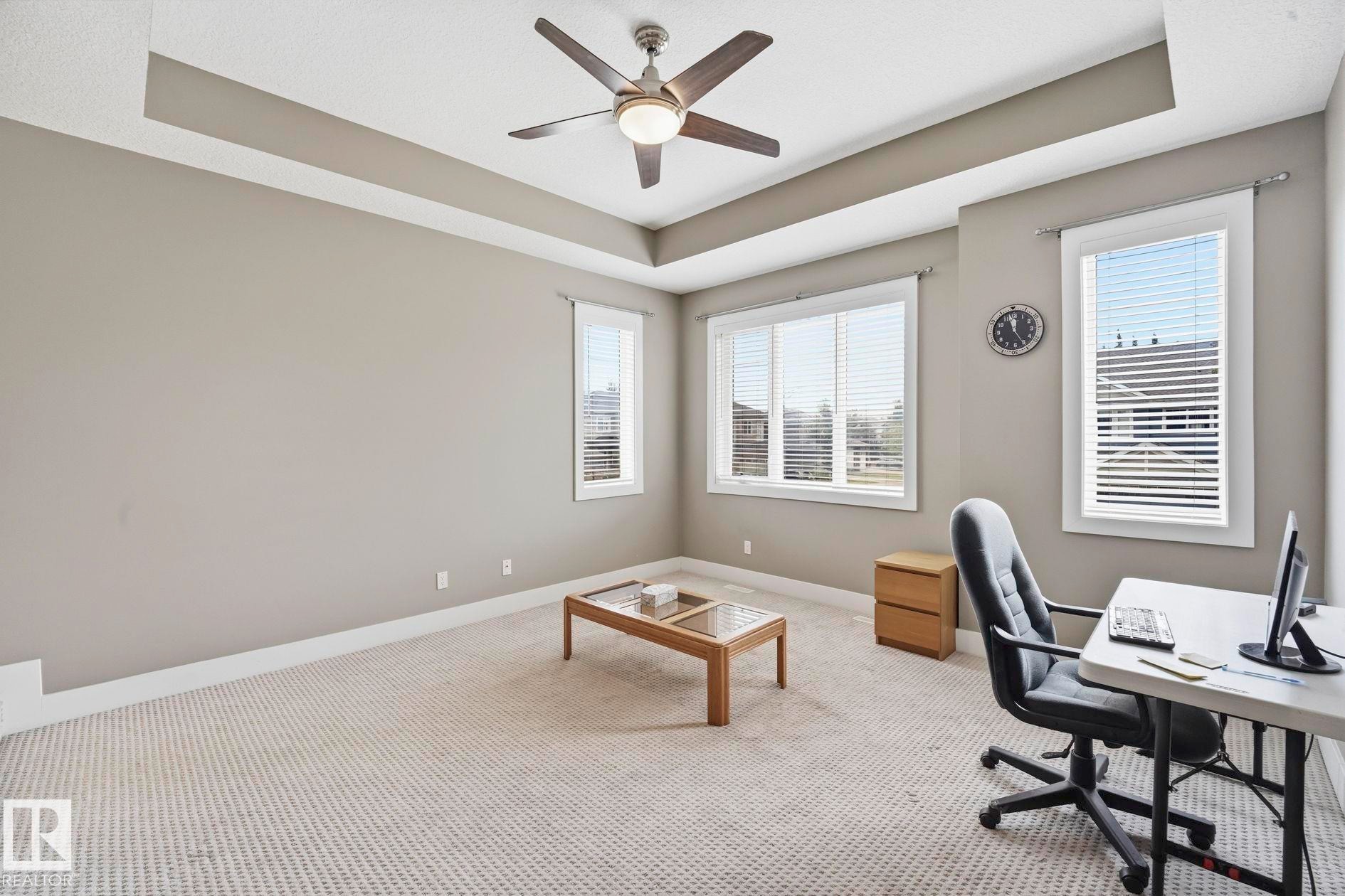Spacious room featuring a tray ceiling with a ceiling fan, abundant natural light from multiple windows with blinds, and neutral carpet flooring - 1051 174 Street Sw, Edmonton, AB - Indoor Photo Showing Office