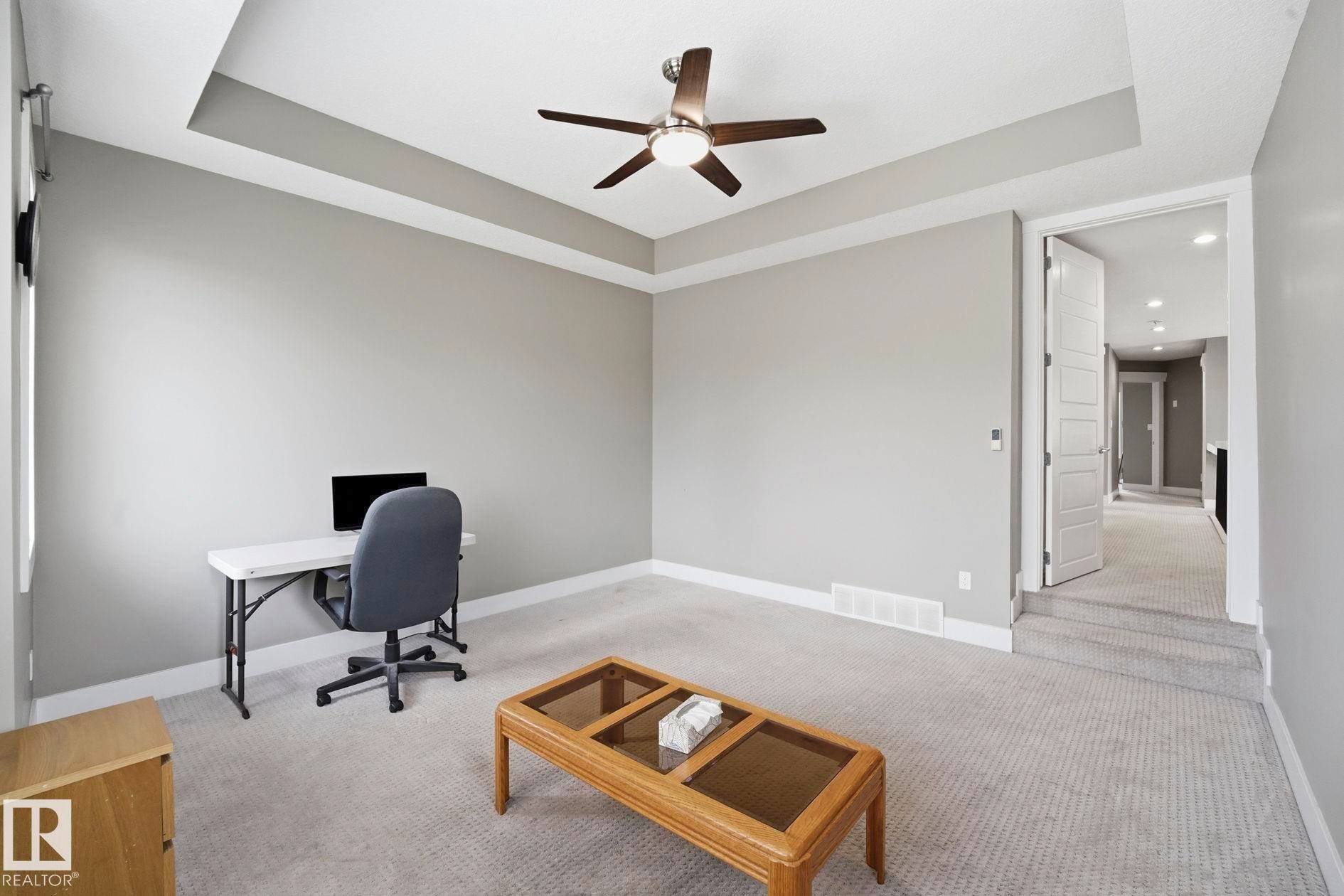 Spacious room featuring a tray ceiling, ceiling fan with integrated lighting, light gray wall finishes, white baseboards, and neutral-toned carpeting - 1051 174 Street Sw, Edmonton, AB - Indoor