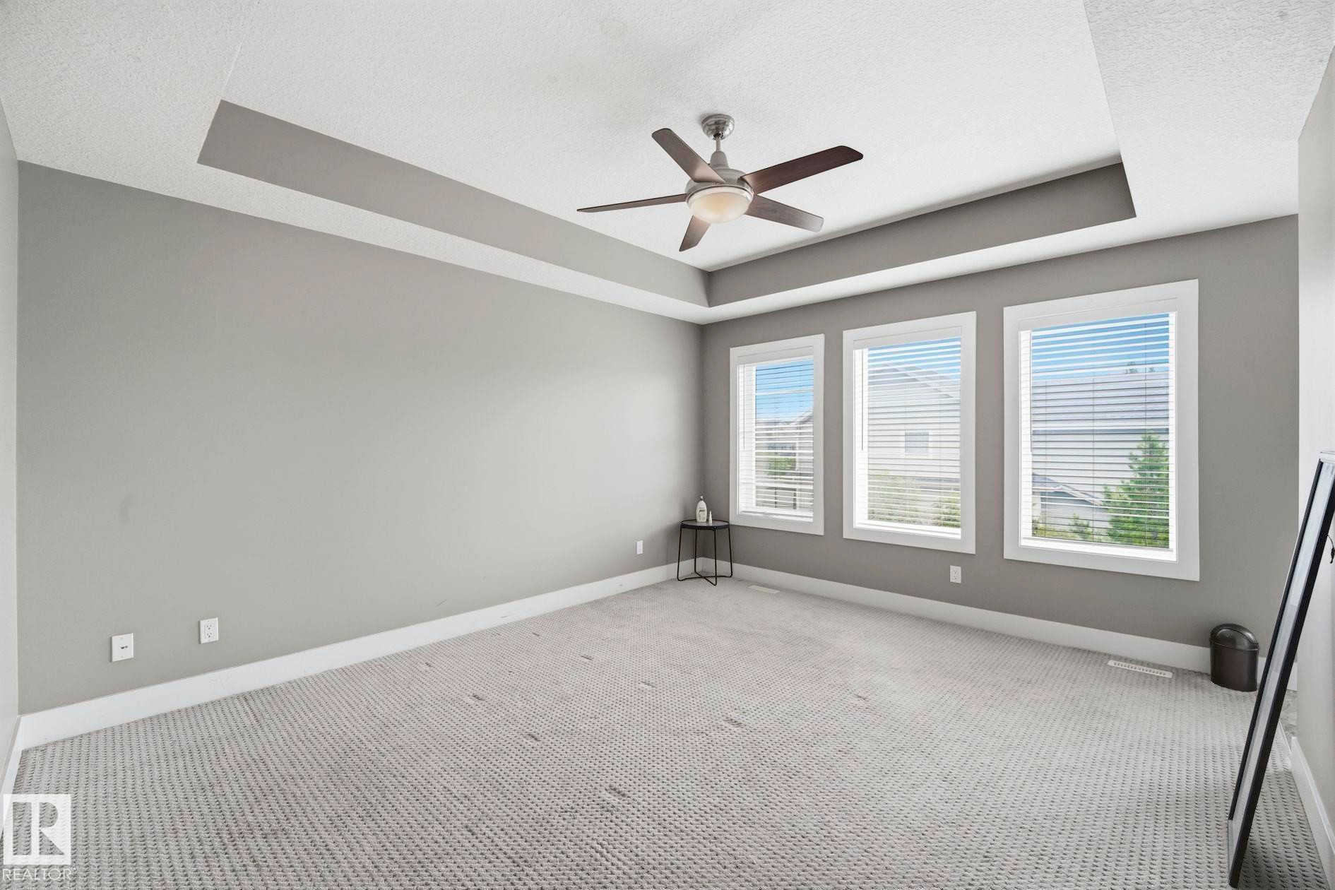 Spacious room featuring a tray ceiling, ceiling fan with integrated lighting, and three large windows with white trim - 1051 174 Street Sw, Edmonton, AB - Indoor Photo Showing Other Room