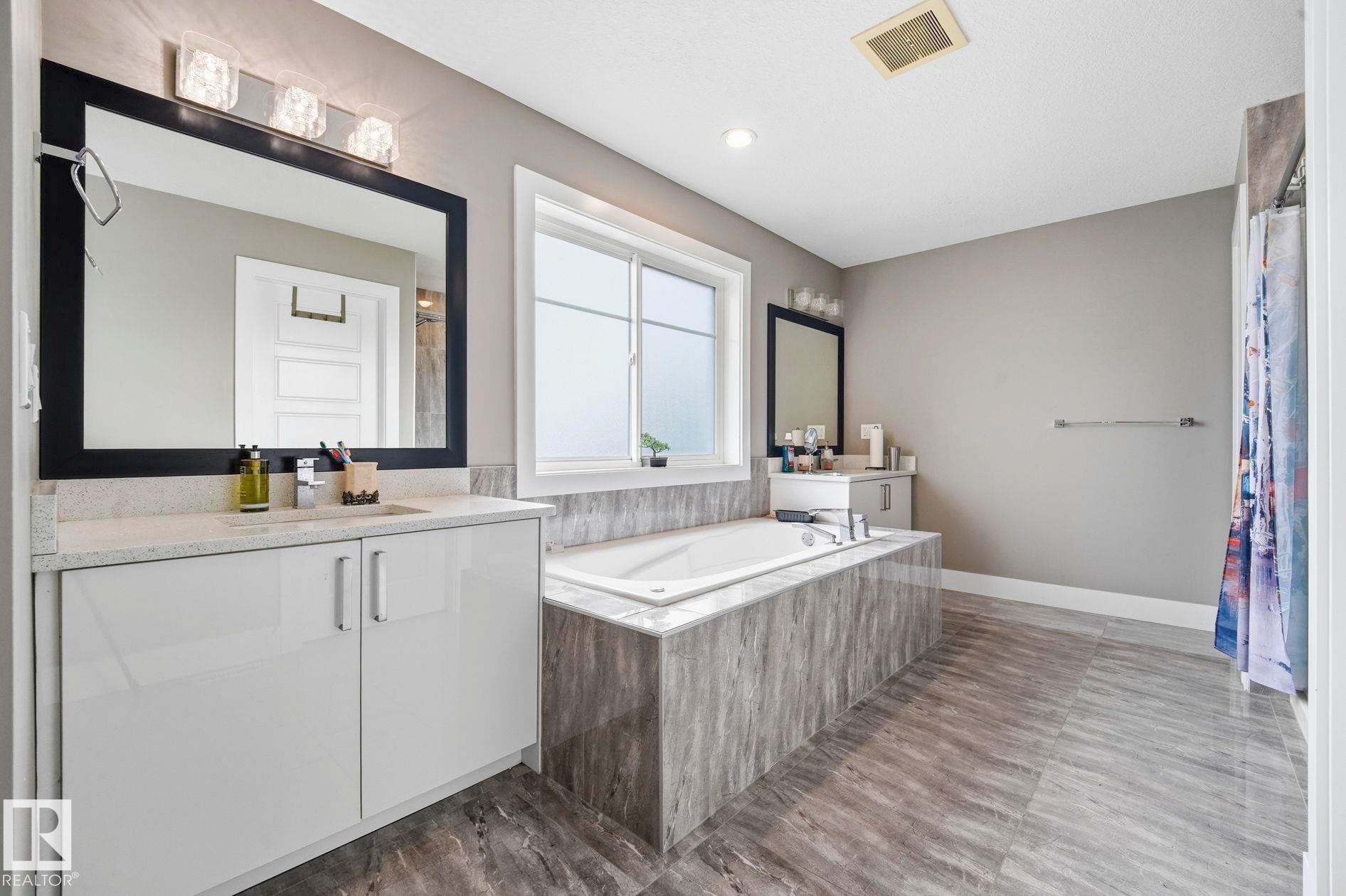 Bathroom featuring a built-in soaking tub with a tiled surround, dual vanity with light countertops, contemporary cabinetry, and large format floor tiling - 1051 174 Street Sw, Edmonton, AB - Indoor Photo Showing Bathroom