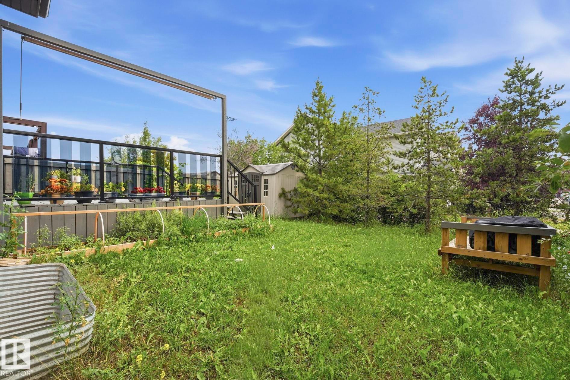 Expansive backyard featuring a multi-level deck with glass panel railing, a built-in garden bed, mature trees, and a storage shed - 1051 174 Street Sw, Edmonton, AB - Outdoor