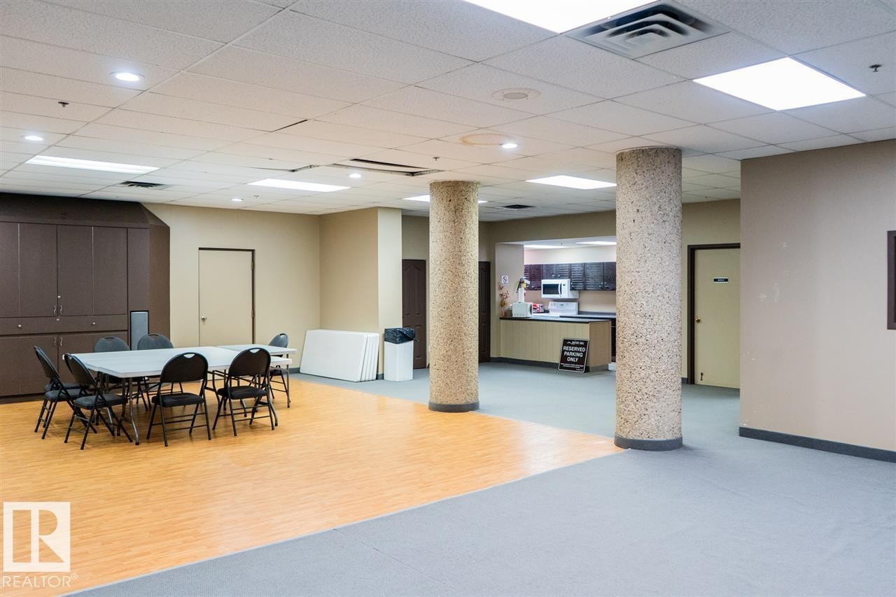 Spacious common area featuring mixed flooring with wood-finish sections and carpeting, two textured support columns, and a dedicated kitchenette with dark cabinetry and white countertops - #509 10149 Saskatchewan Dr Nw, Edmonton, AB - Indoor