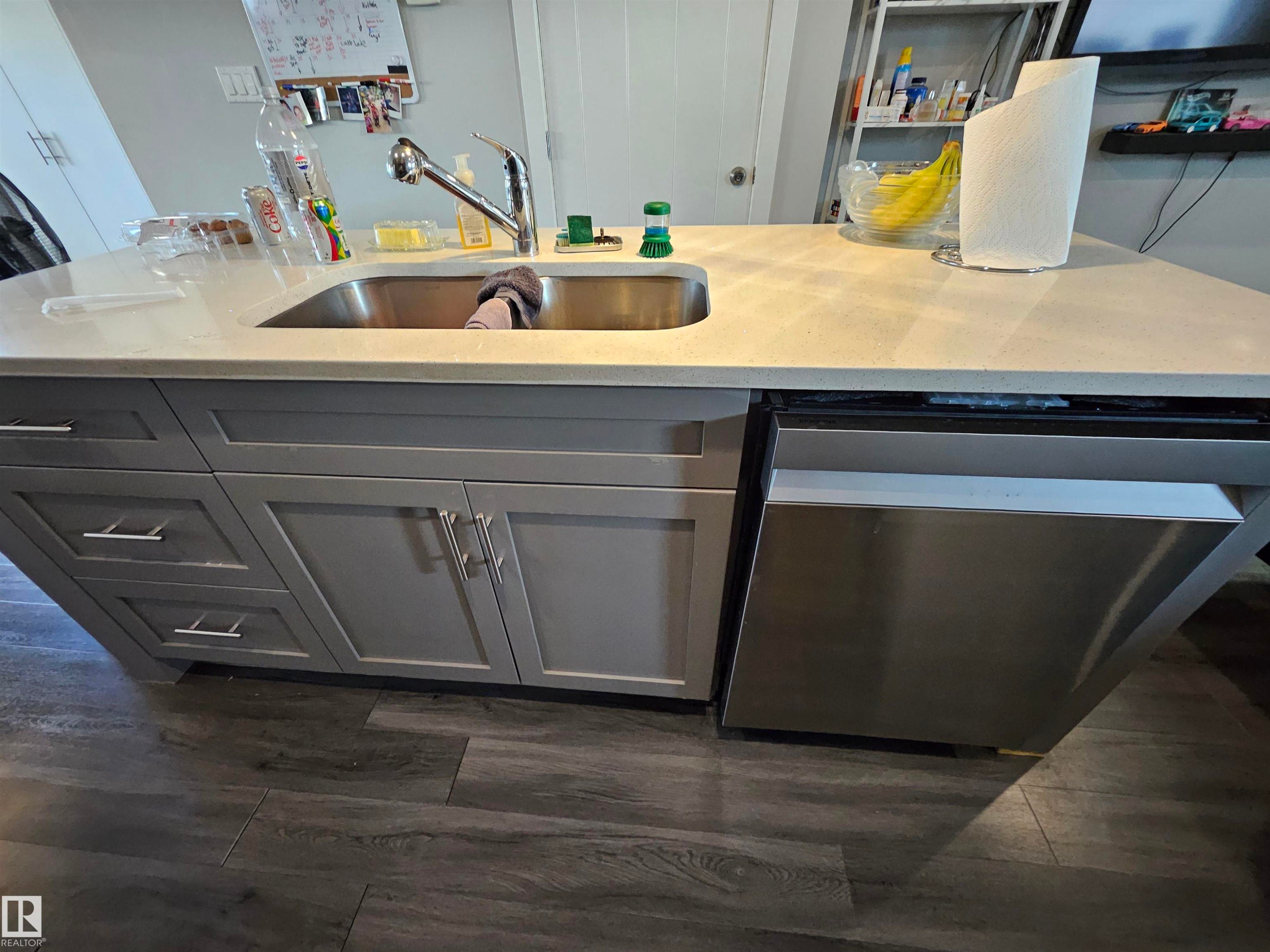 Kitchen island featuring a stainless steel sink, a pull-down faucet, light-toned countertop, gray shaker-style cabinetry with silver hardware, and an integrated stainless steel dishwasher - 13309 104 Street Nw, Edmonton, AB - Indoor Photo Showing Other Room