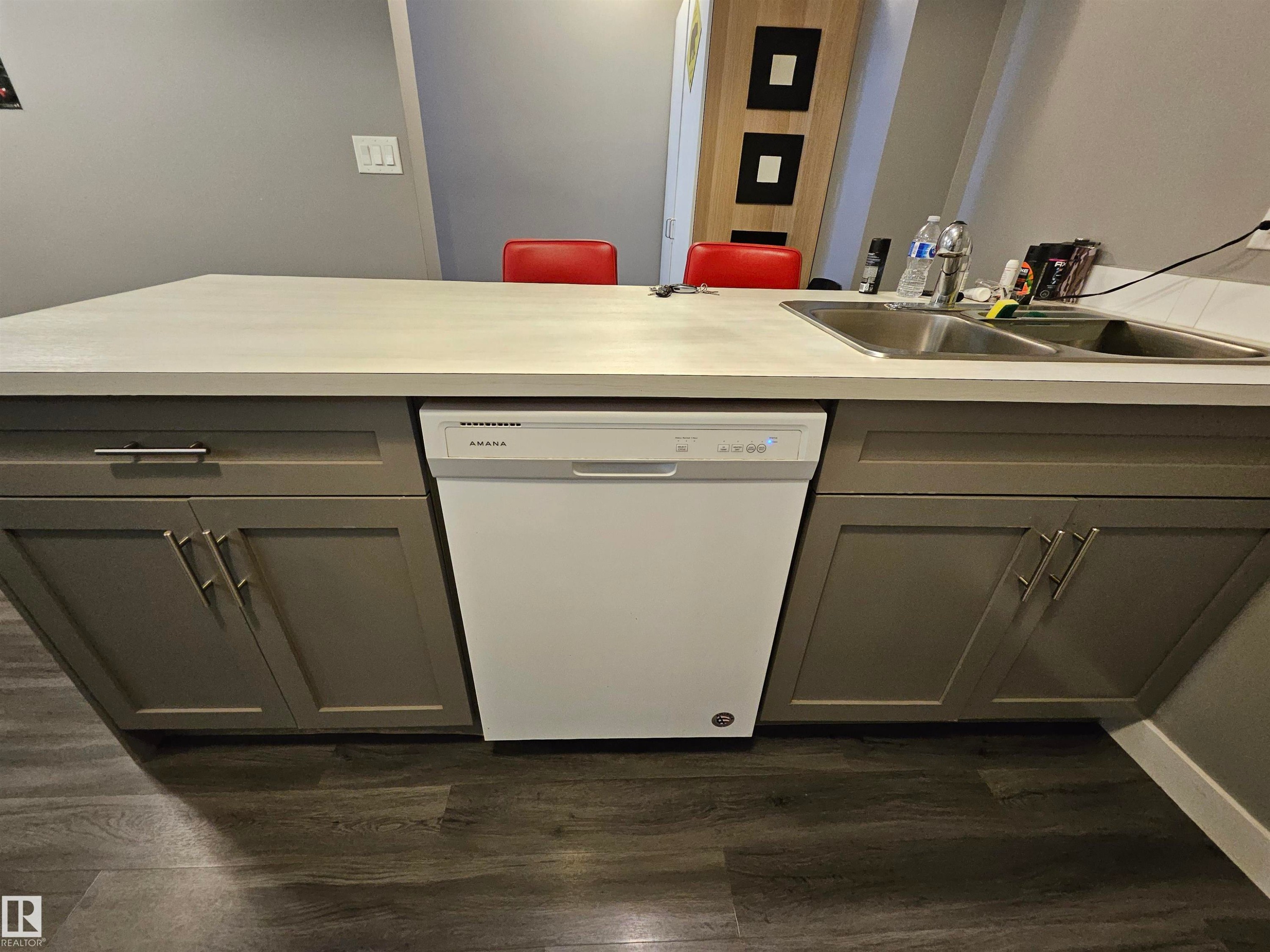 Kitchen island featuring a double basin stainless steel sink, a white Amana dishwasher, and gray shaker-style cabinetry with silver hardware, all complemented by wood-finish flooring - 13309 104 Street Nw, Edmonton, AB - Indoor Photo Showing Other Room