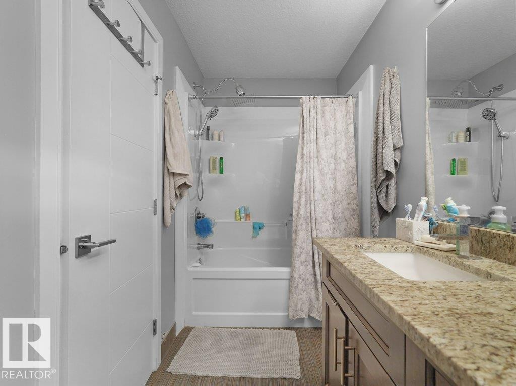 Bathroom featuring a shower-tub combination with built-in shelving, a single vanity with an integrated sink, a stone-finish countertop, and wood-finish flooring - 7909 105 Avenue Nw, Edmonton, AB - Indoor Photo Showing Bathroom