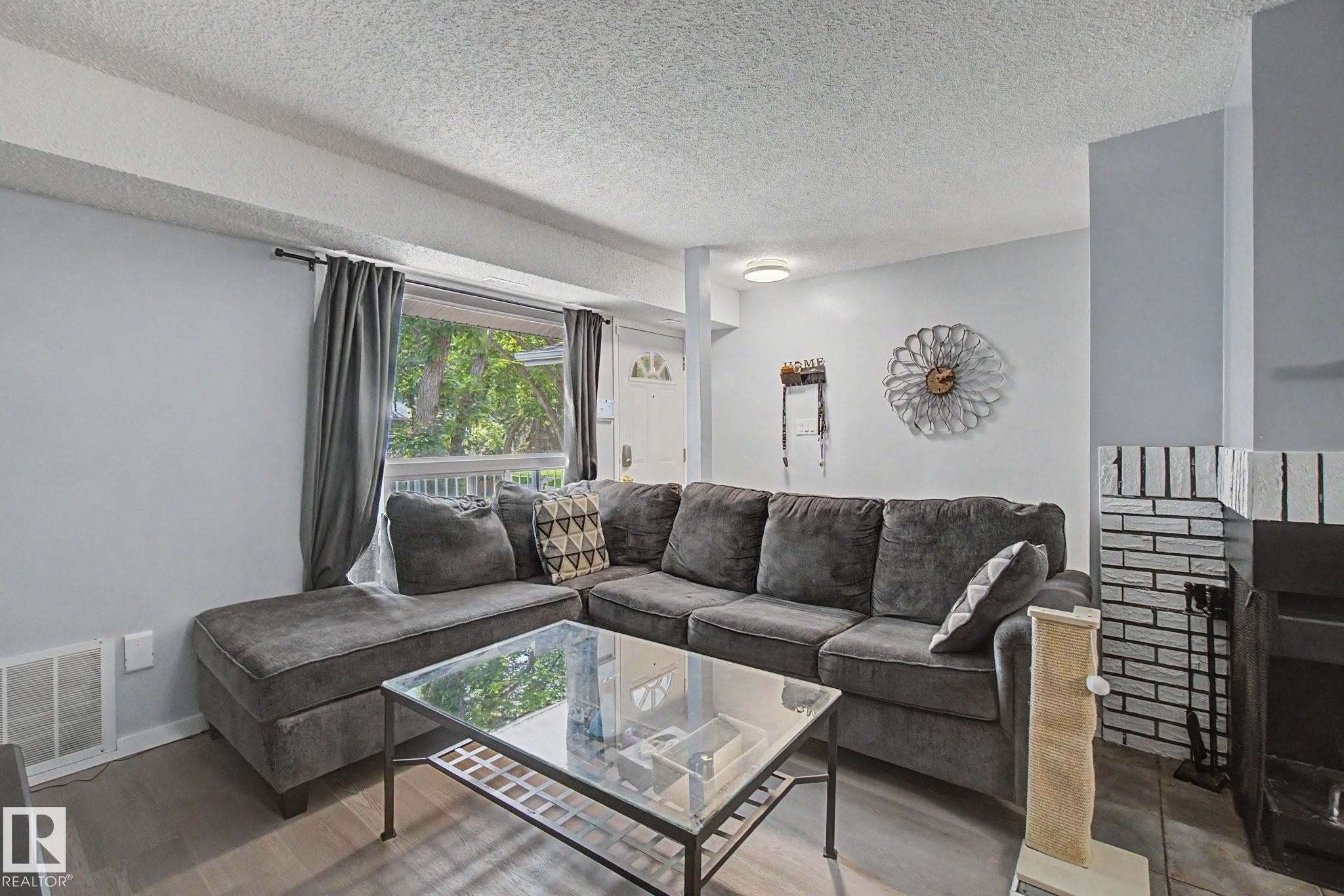 Bright living area featuring light gray walls, wood-finish flooring, and a large window with outdoor greenery - #39 3111 142 Av Nw, Edmonton, AB - Indoor Photo Showing Living Room