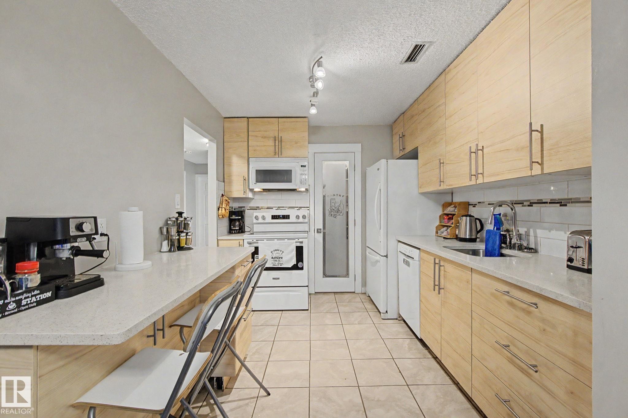 Light wood-finish cabinetry with brushed nickel hardware, white quartz countertops, full-size white appliances, tile flooring, and a glass-paneled pantry door - #39 3111 142 Av Nw, Edmonton, AB - Indoor Photo Showing Kitchen